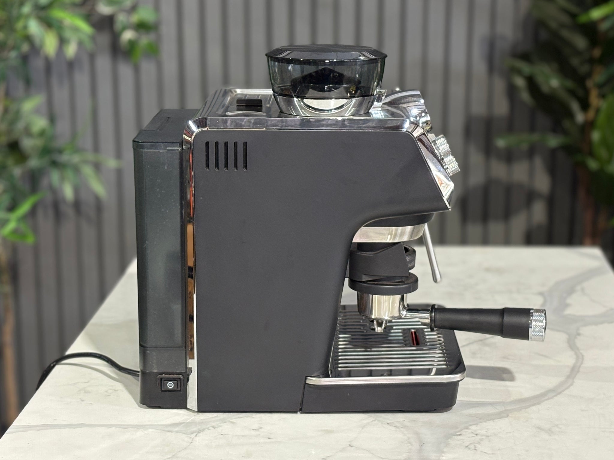 A Delonghi La Specialista Arte 1 Group Coffee Machine in Black sitting on a marble bench. There is greenery in the background. The image is taken from the side of the machine.