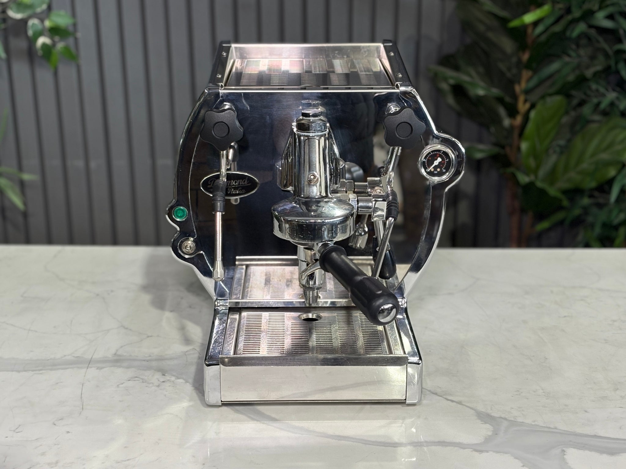 A Diamond Cuadra 1 Group Espresso Coffee Machine with Stainless finish sitting on a marble bench. There is greenery in the background. The image is taken from the front of the machine but at a high angle so that you can see the front of the machine and also the top of the machine.