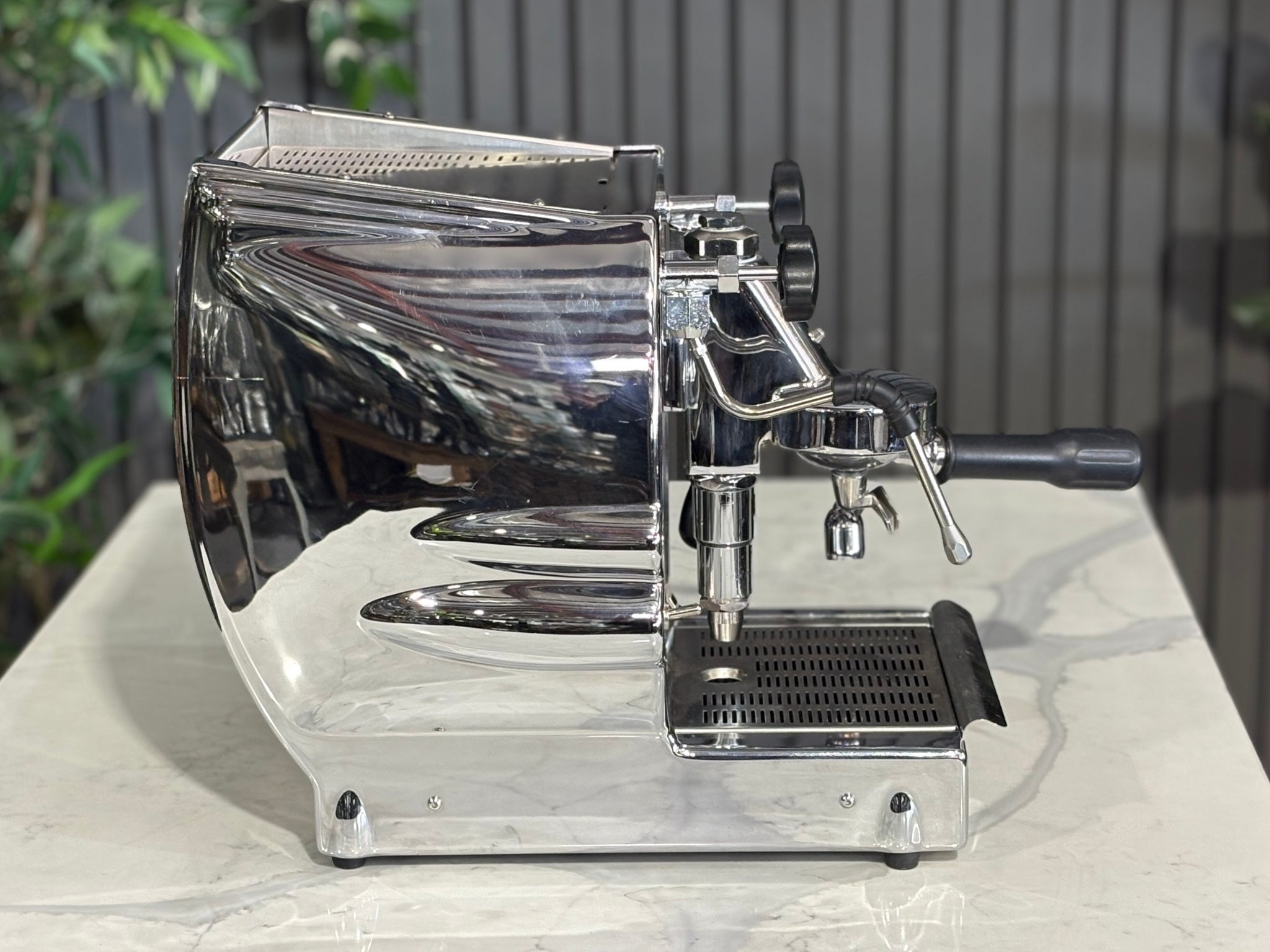 A Diamond Cuadra 1 Group Espresso Coffee Machine with Stainless finish sitting on a marble bench. There is greenery in the background. The image is taken from the side of the machine.