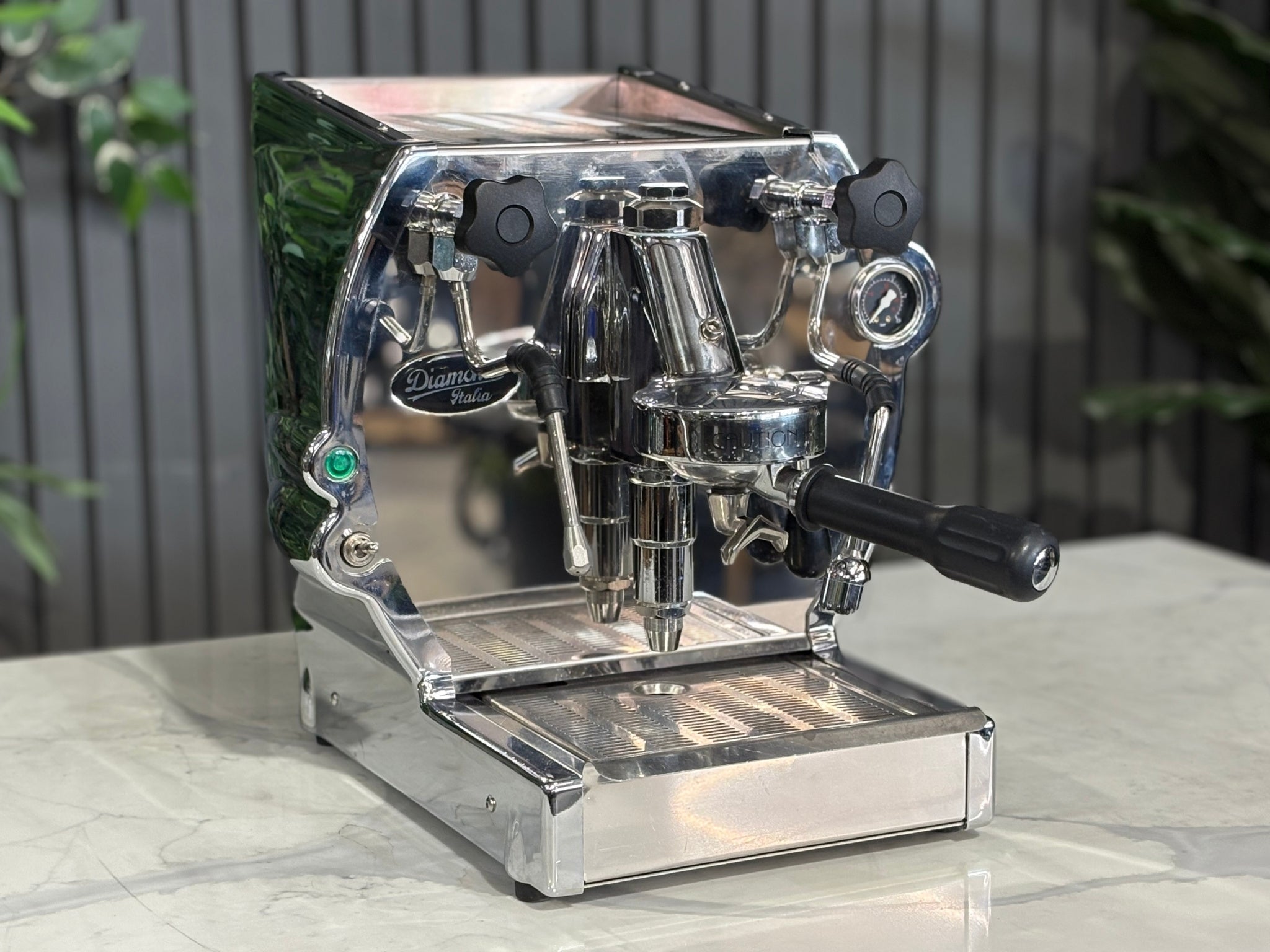 A Diamond Cuadra 1 Group Espresso Coffee Machine with Stainless finish is sitting on a marble bench. There is greenery in the background. The image is taken from the front of the machine and the machine is sitting on an angle so that you can see the front of the machine and the side.