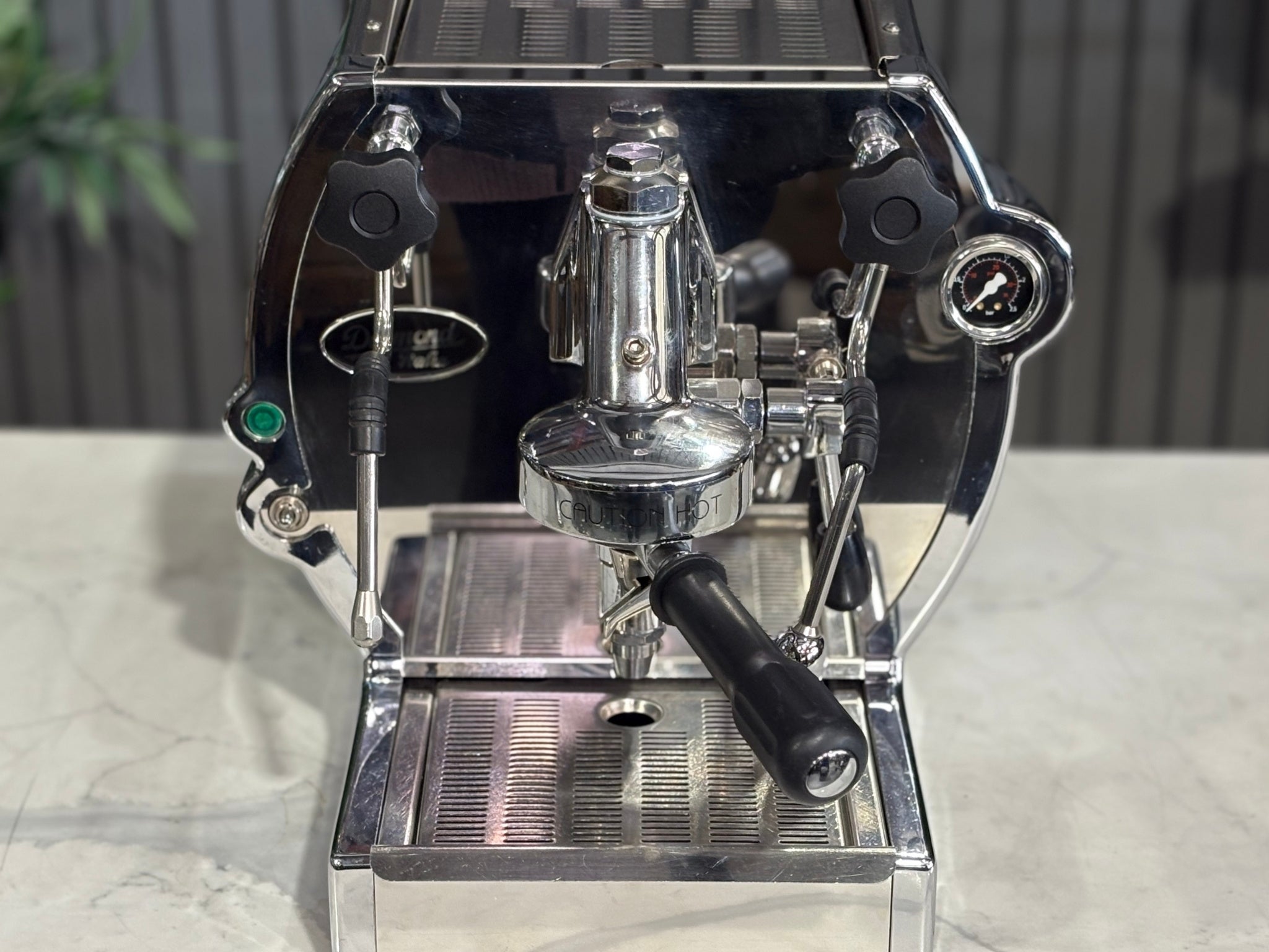 A Diamond Cuadra 1 Group Espresso Coffee Machine with Stainless finish
is sitting on a marble bench. There is greenery in the background. The image is a close up image taken from the front of the machine  so that you can see the front of the machine, the controls including buttons, lights and group handle.