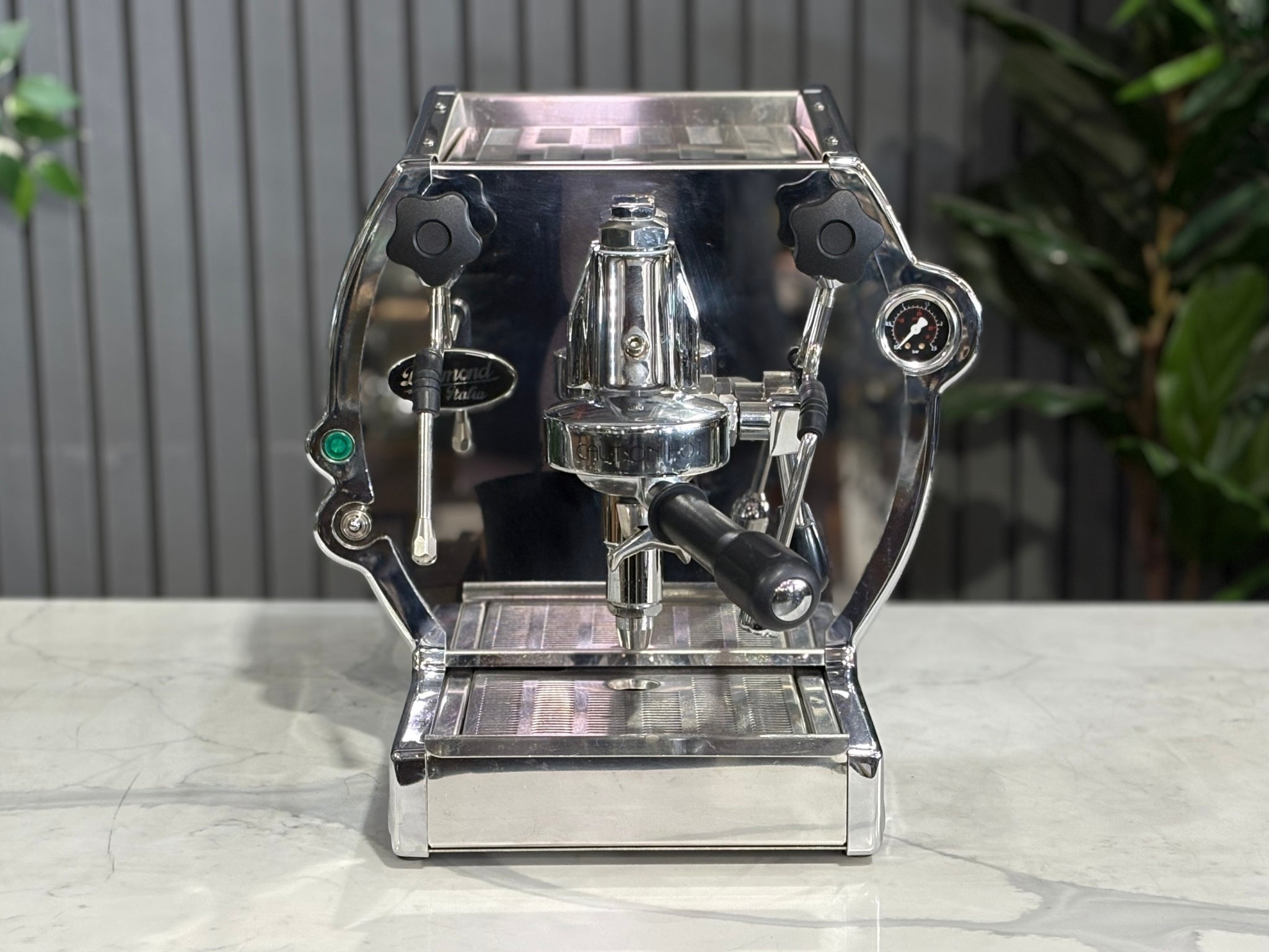 A Diamond Cuadra 1 Group Espresso Coffee Machine with Stainless finish sitting on a marble bench. There is greenery in the background. The image is taken from the front of the machine so that you can see the front of the machine, control panel, buttons and group heads.