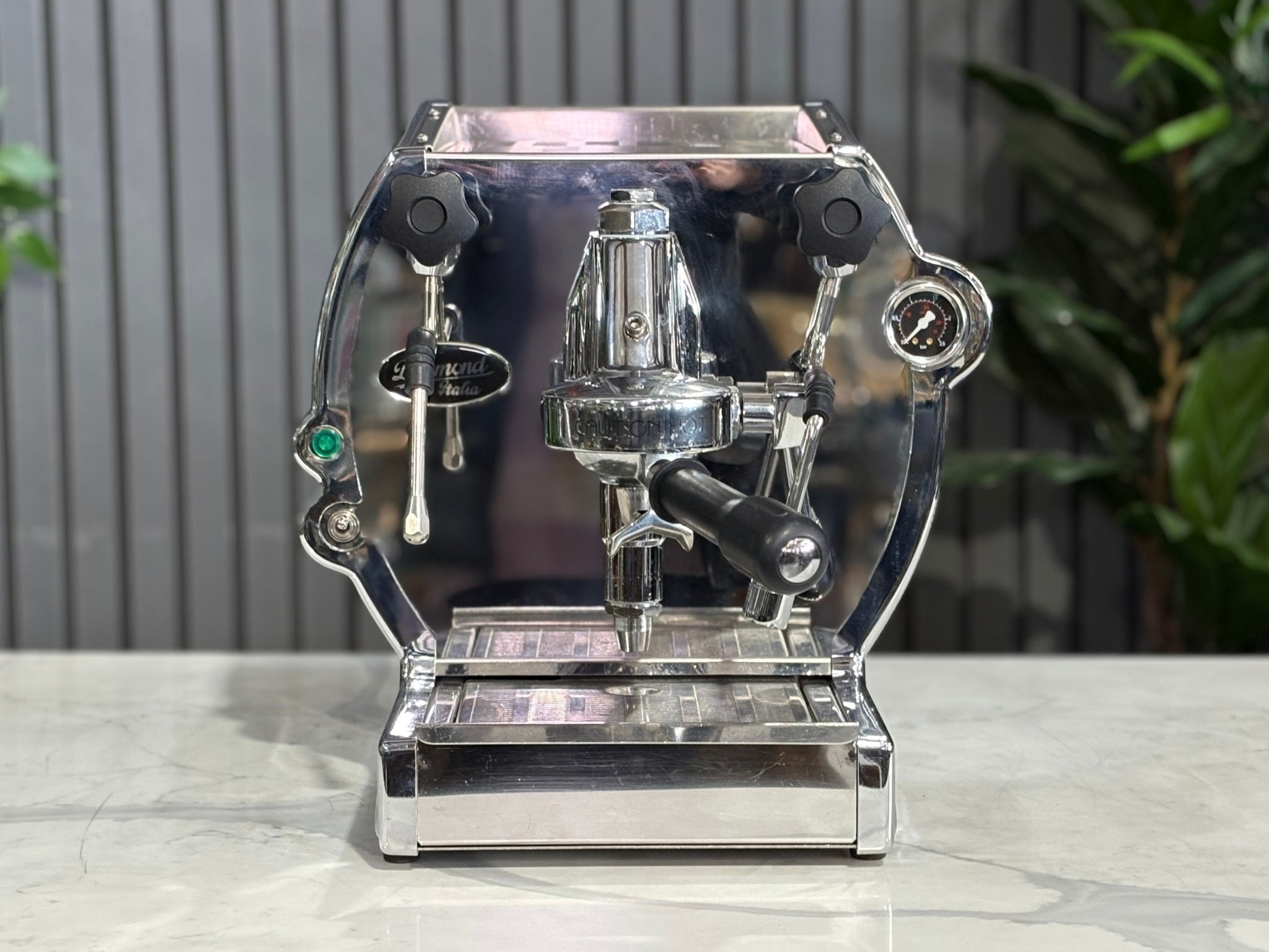 A Diamond Cuadra 1 Group Espresso Coffee Machine with Stainless finish sitting on a marble bench. There is greenery in the background. The image is taken from the front of the machine so that you can see the front of the machine, control panel, buttons and group heads.