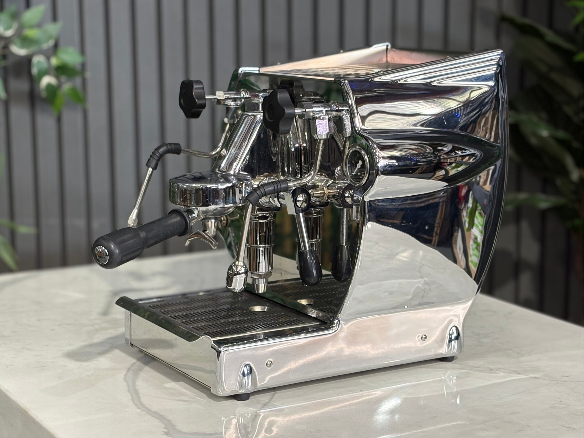 A Diamond Cuadra 1 Group Espresso Coffee Machine with Stainless finish sitting on a marble bench. There is greenery in the background. The image is taken from the side of the machine.