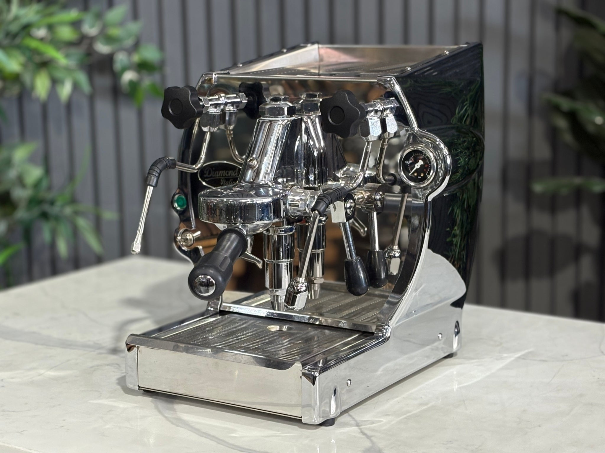 A Diamond Cuadra 1 Group Espresso Coffee Machine with Stainless finish is sitting on a marble bench. There is greenery in the background. The image is taken from the front of the machine and the machine is sitting on an angle so that you can see the front of the machine and the side.