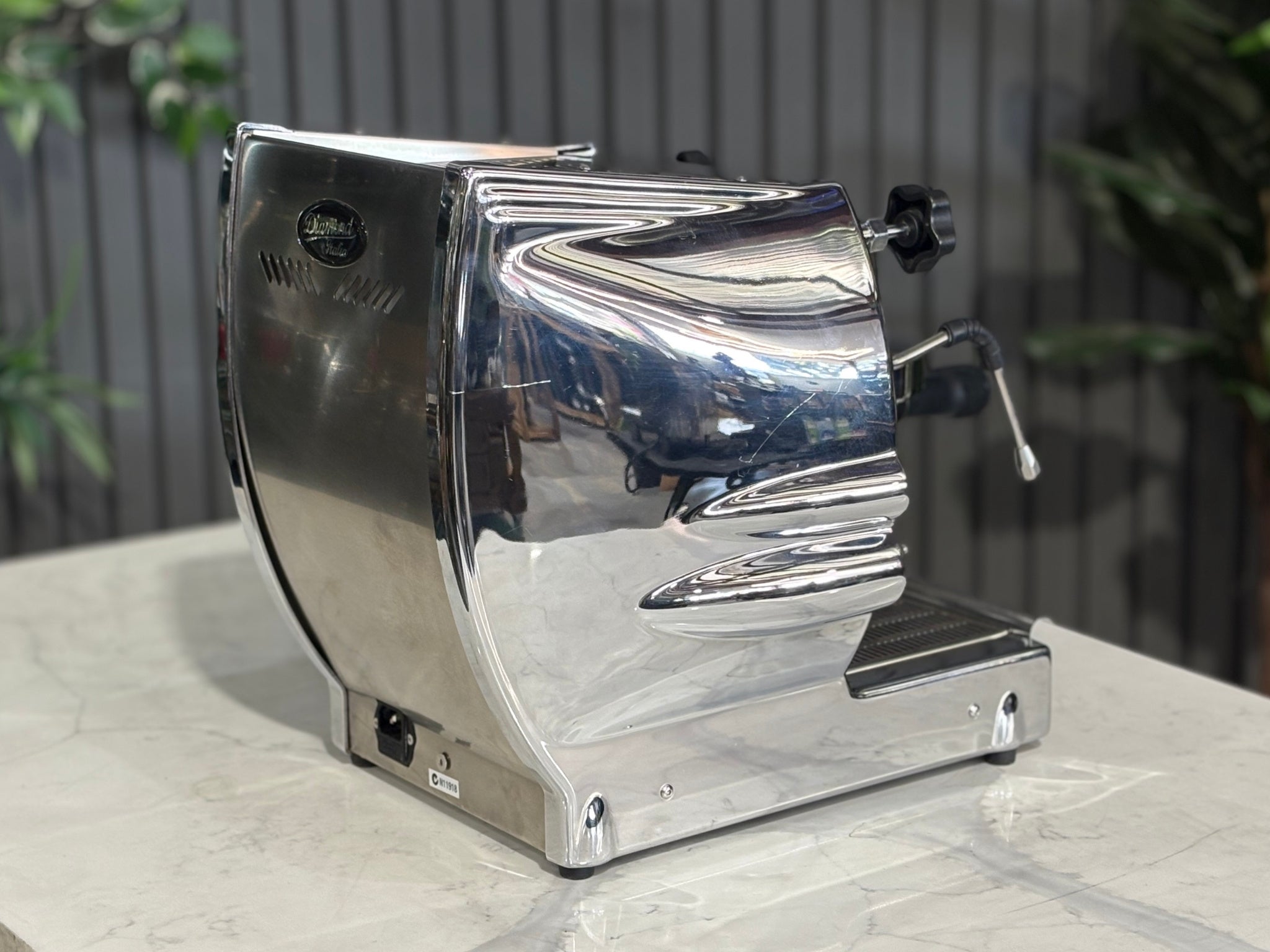 A Diamond Cuadra 1 Group Espresso Coffee Machine with Stainless finish itting on a marble bench. There is greenery in the background. The image is taken from behind the machine and the machine is sitting on an angle so that you can see the back of the machine and the side.