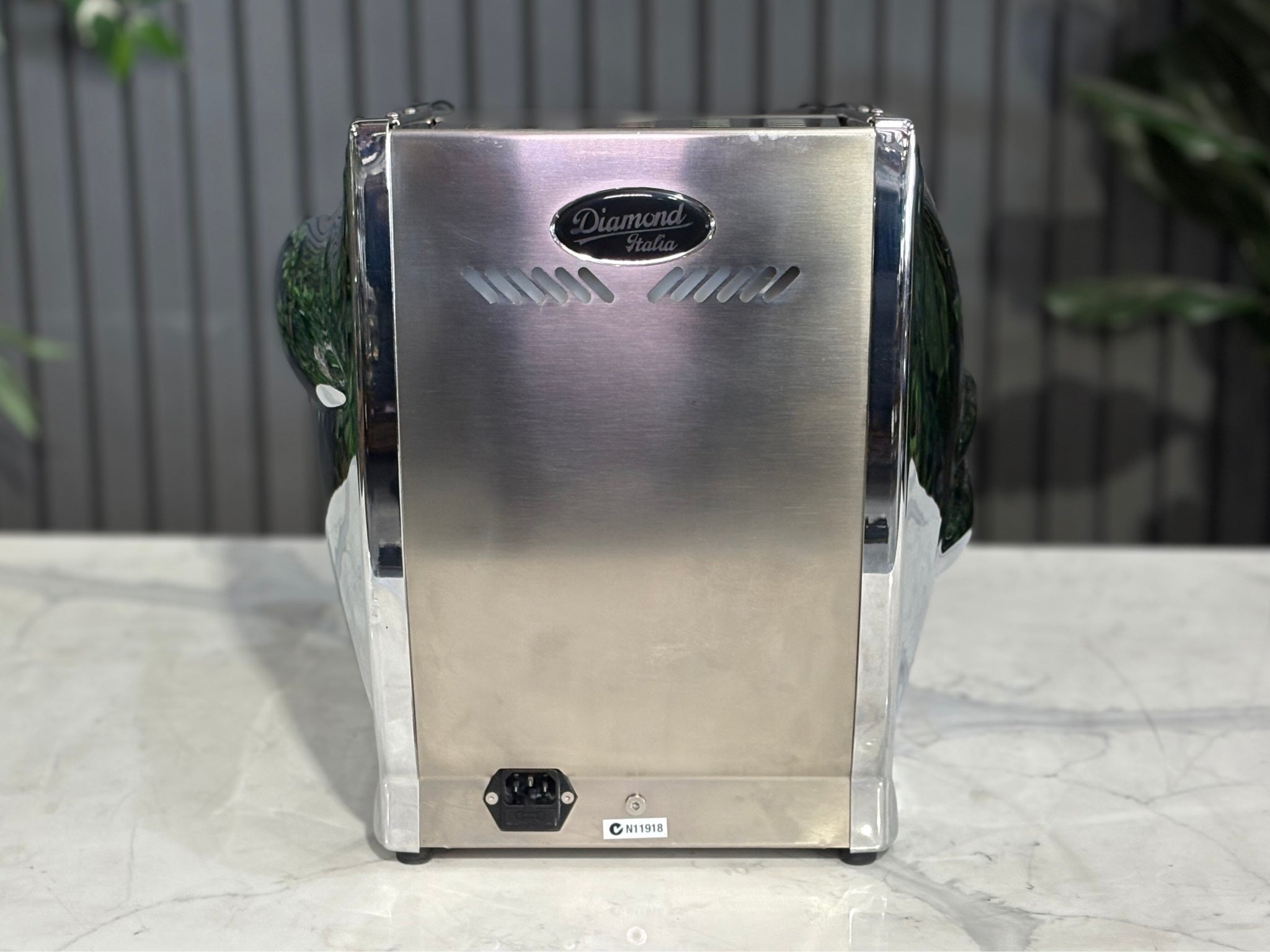 A Diamond Cuadra 1 Group Espresso Coffee Machine with Stainless finish s sitting on a marble bench. There is greenery in the background. The image is taken from behind the machine so the back is visible.