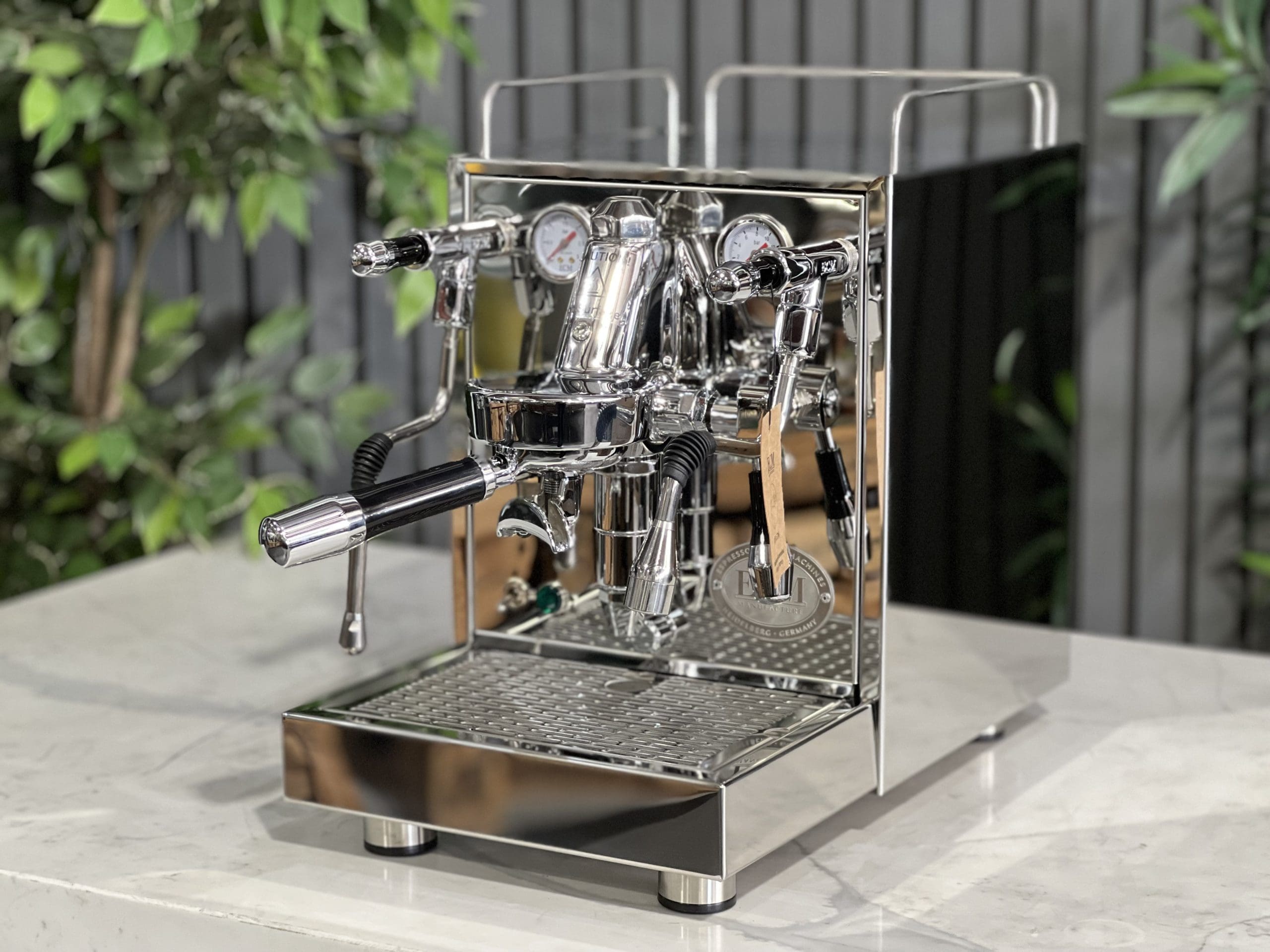 ECM Mechanika Max Rotary 1 Group Espresso Coffee Machine Stainless - New shown at a front angle on a white marble bench with greenery in the background, highlighting its polished stainless steel body, flick taps, E61 group head, and cup warming tray.