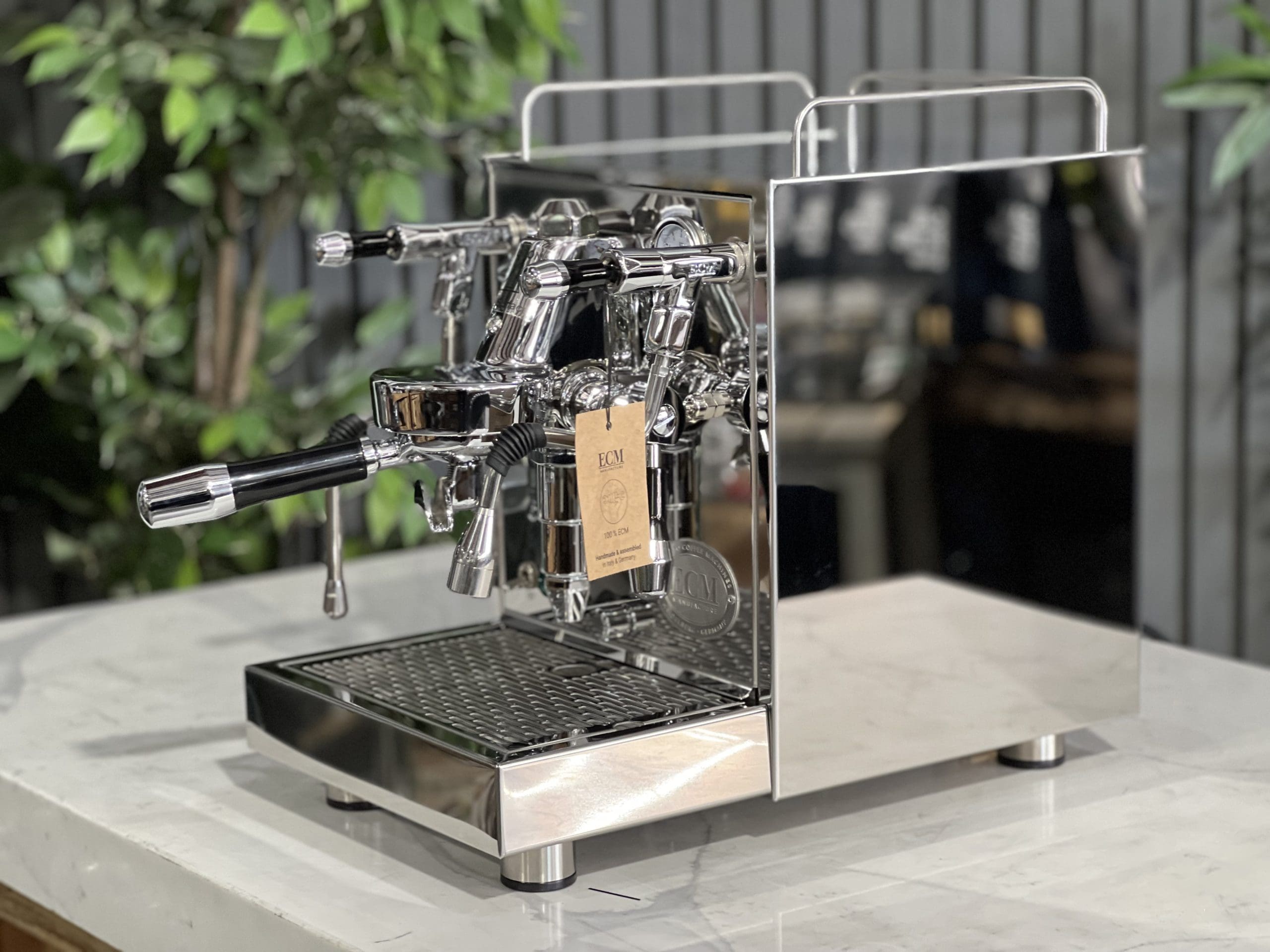 Side profile of the ECM Mechanika Max Rotary 1 Group Espresso Coffee Machine Stainless - New on a white marble bench with greenery behind, displaying its polished stainless steel side panel, steam wand, and rotary pump design.