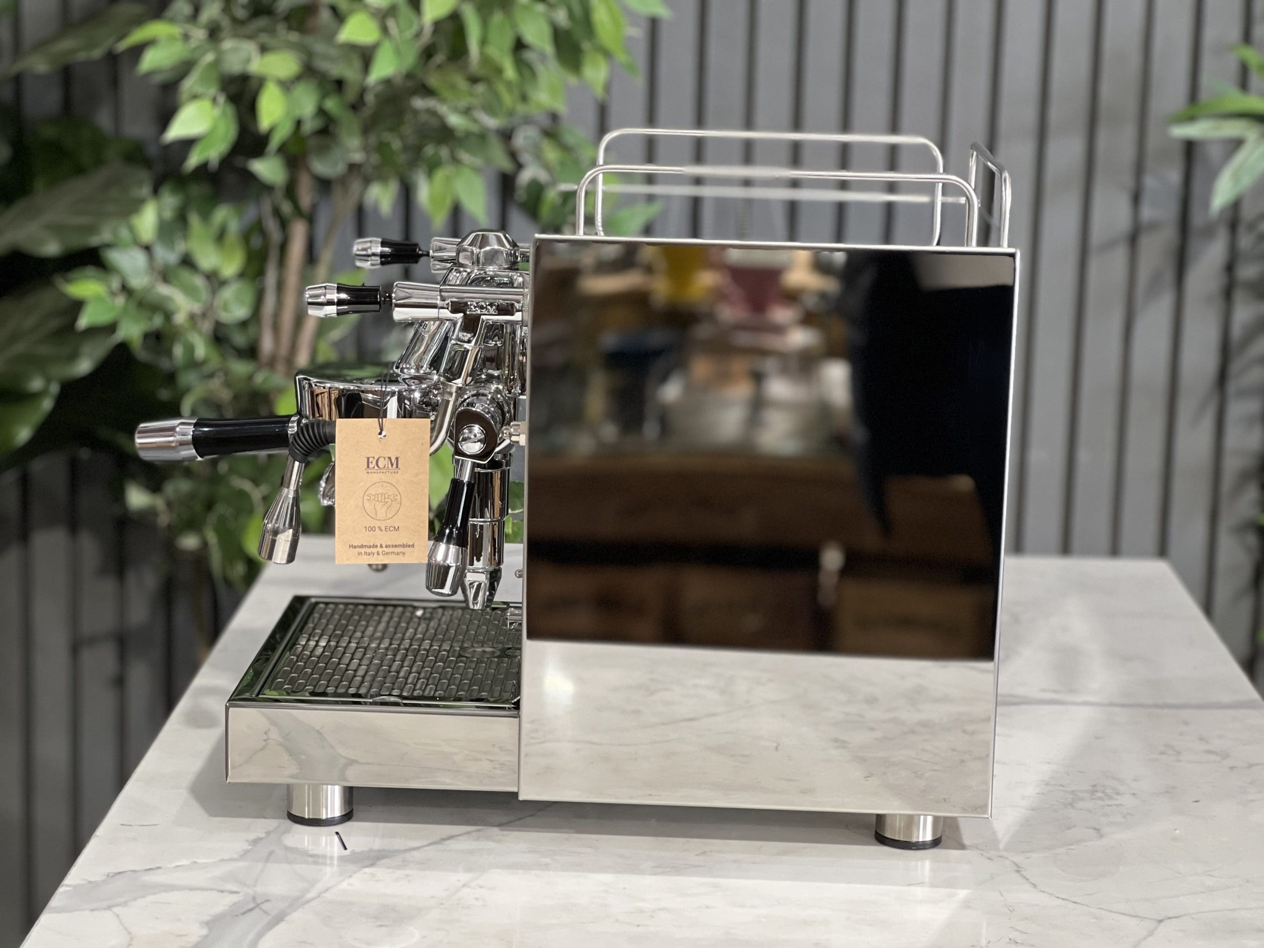 Side profile of the ECM Mechanika Max Rotary 1 Group Espresso Coffee Machine Stainless - New on a white marble bench with greenery behind, displaying its polished stainless steel side panel, steam wand, and rotary pump design.