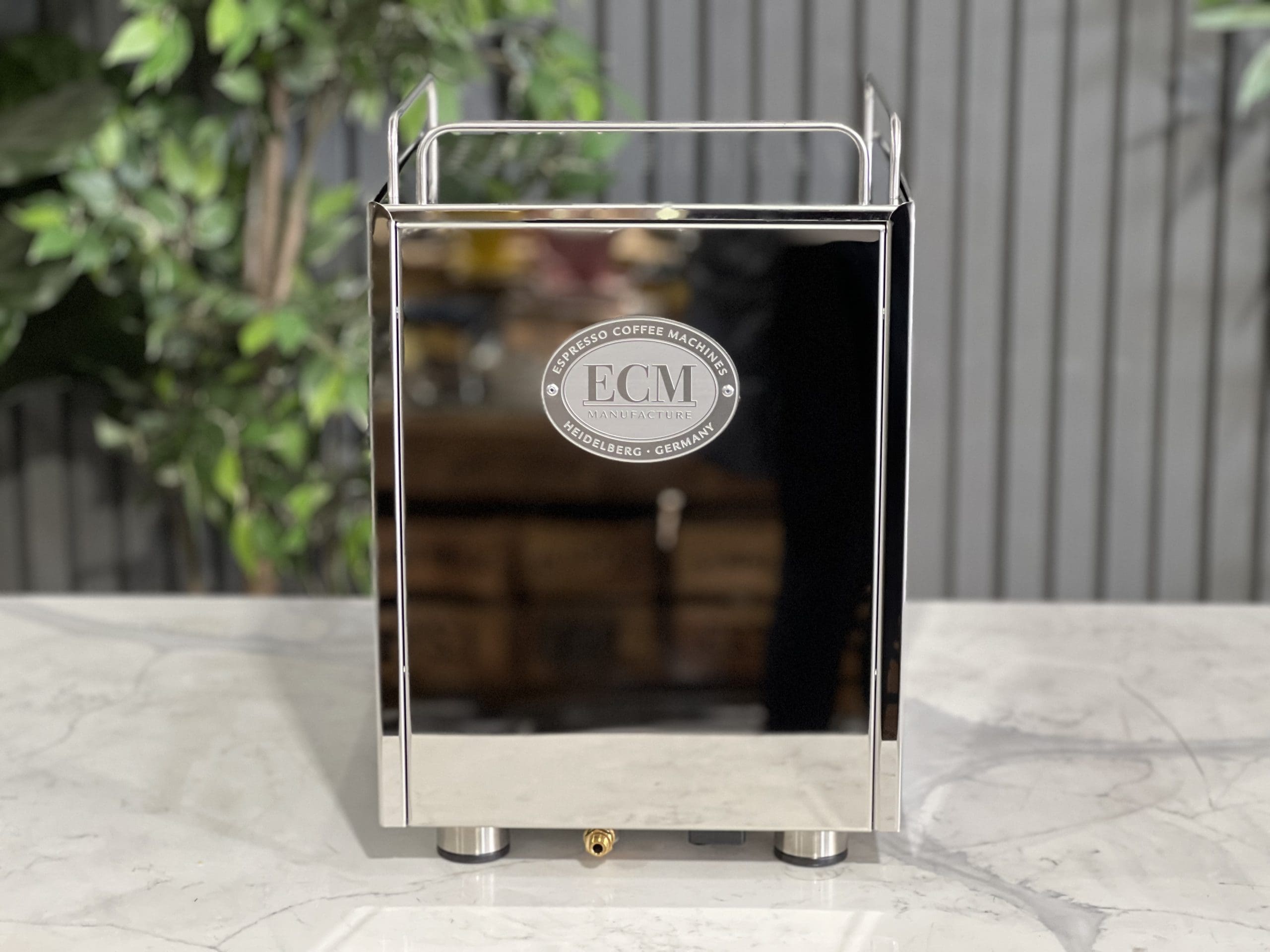 Rear view of the ECM Mechanika Max Rotary 1 Group Espresso Coffee Machine Stainless - New, highlighting the stainless steel back panel, ventilation detailing, and power connection points, set on a marble bench with greenery behind.