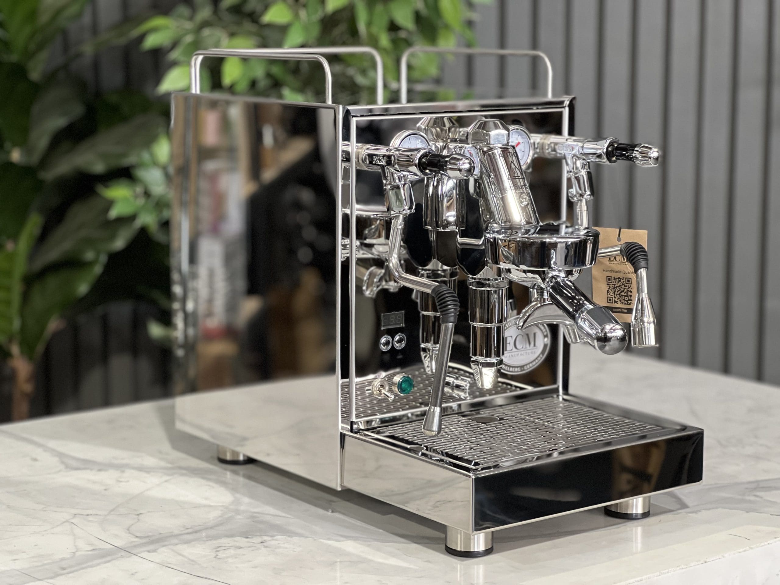 ECM Mechanika Max Rotary 1 Group Espresso Coffee Machine Stainless - New shown at a front angle on a white marble bench with greenery in the background, highlighting its polished stainless steel body, flick taps, E61 group head, and cup warming tray.