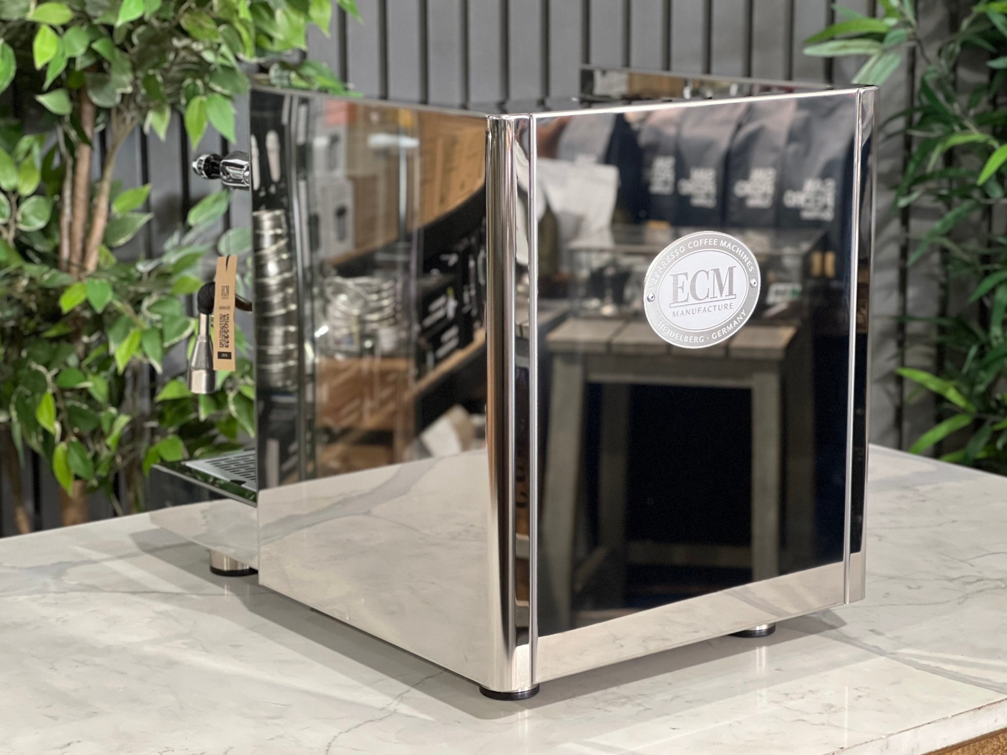 ECM Technika V Profi Rotary PID 1 Group Coffee Machine Stainless – New espresso coffee machine angled from the back, showing the stainless steel rear panel, top cup tray, and part of the E61 group head on a marble bench with greenery behind.