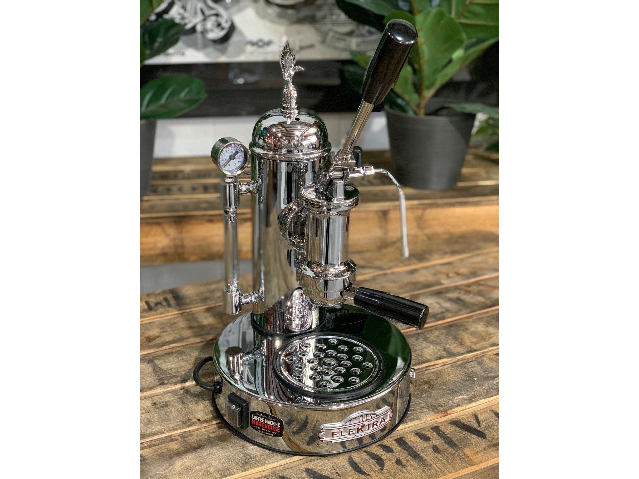A brand new Elektra Micro Casa Leva 1 Group Coffee Machine with Chrome finish is sitting on a wooden bench. There is greenery in the background. The image is taken from the front of the machine and the machine is sitting on an angle so that you can see the front of the machine and the side.