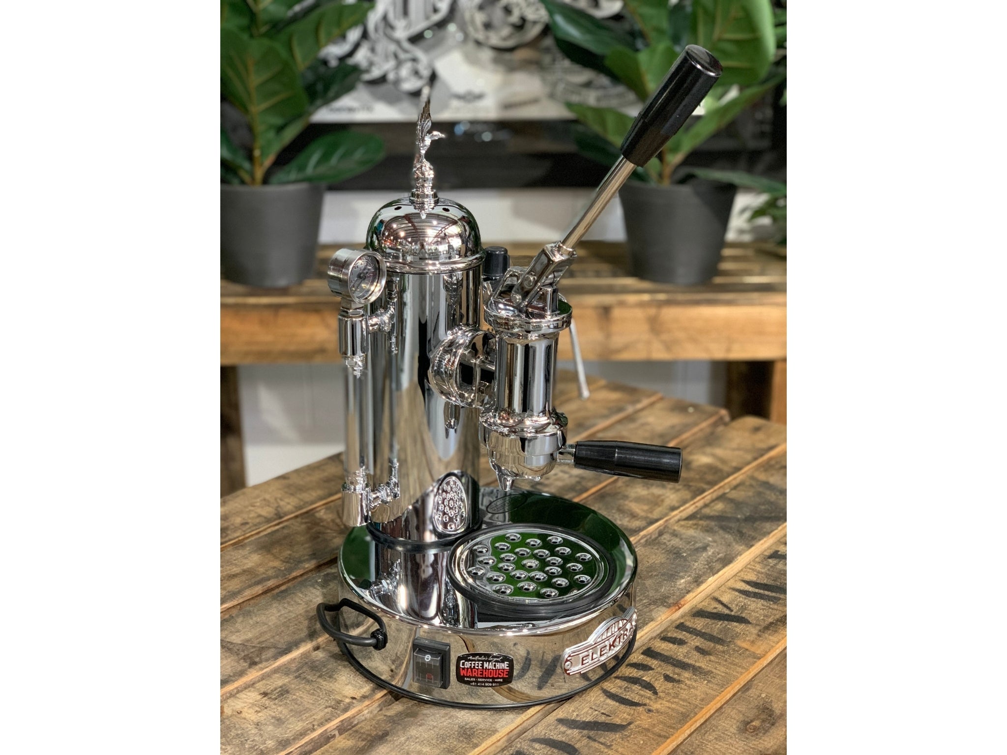 A brand new Elektra Micro Casa Leva 1 Group Coffee Machine with Chrome finish is sitting on a wooden bench. There is greenery in the background. The image is taken from the front of the machine and the machine is sitting on an angle so that you can see the front of the machine and the side.
