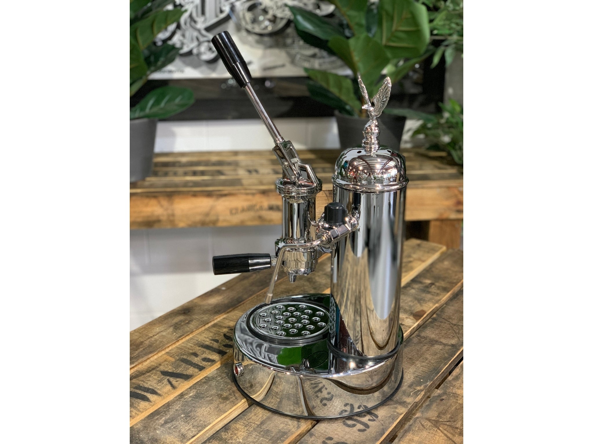 A brand new Elektra Micro Casa Leva 1 Group Coffee Machine with Chrome finish sitting on a wooden bench. There is greenery in the background. The image is taken from the side of the machine.