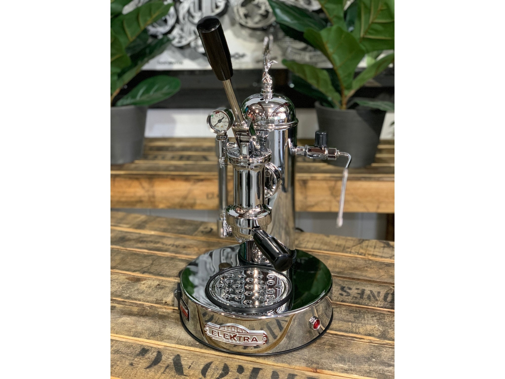 A brand new Elektra Micro Casa Leva 1 Group Coffee Machine with Chrome finish is sitting on a wooden bench. There is greenery in the background. The image is taken from the front of the machine and the machine is sitting on an angle so that you can see the front of the machine and the side.