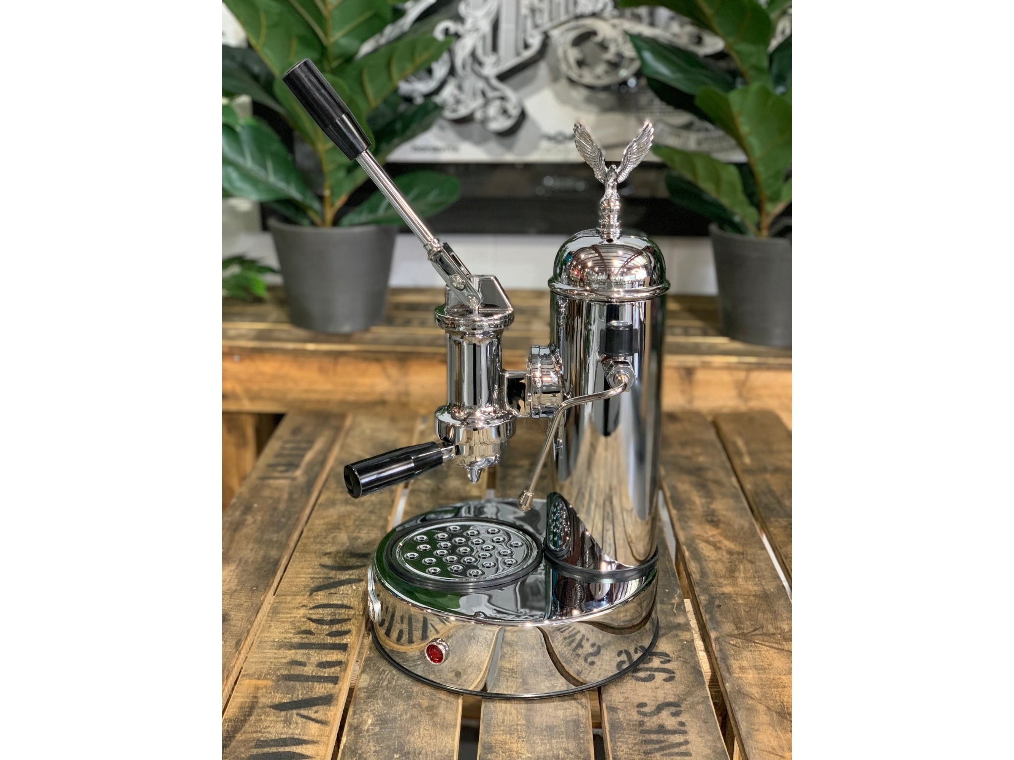 A brand new Elektra Micro Casa Leva 1 Group Coffee Machine with Chrome finish sitting on a wooden bench. There is greenery in the background. The image is taken from the side of the machine.