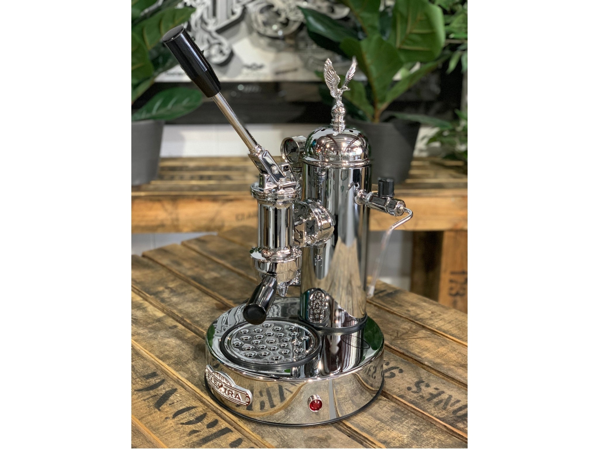 A brand new Elektra Micro Casa Leva 1 Group Coffee Machine with Chrome finish is sitting on a wooden bench. There is greenery in the background. The image is taken from the front of the machine and the machine is sitting on an angle so that you can see the front of the machine and the side.