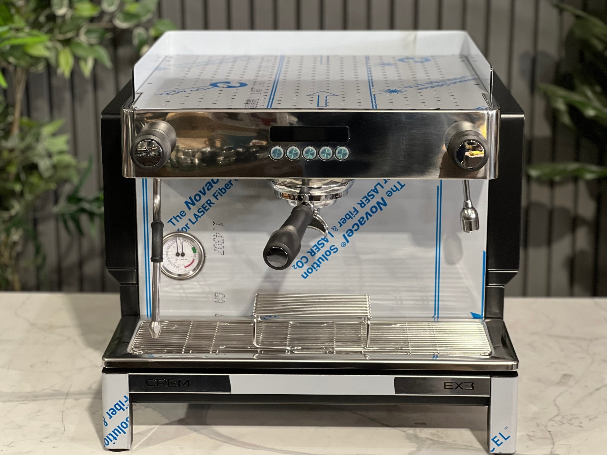 Expobar Crem EX3 Compact 1 Group Coffee Machine Black - New front image from above, showcasing the top panel and cup tray along with the group head and controls. Set on a marble bench with lush greenery in the background, ideal for showcasing this compact espresso coffee machine’s design.