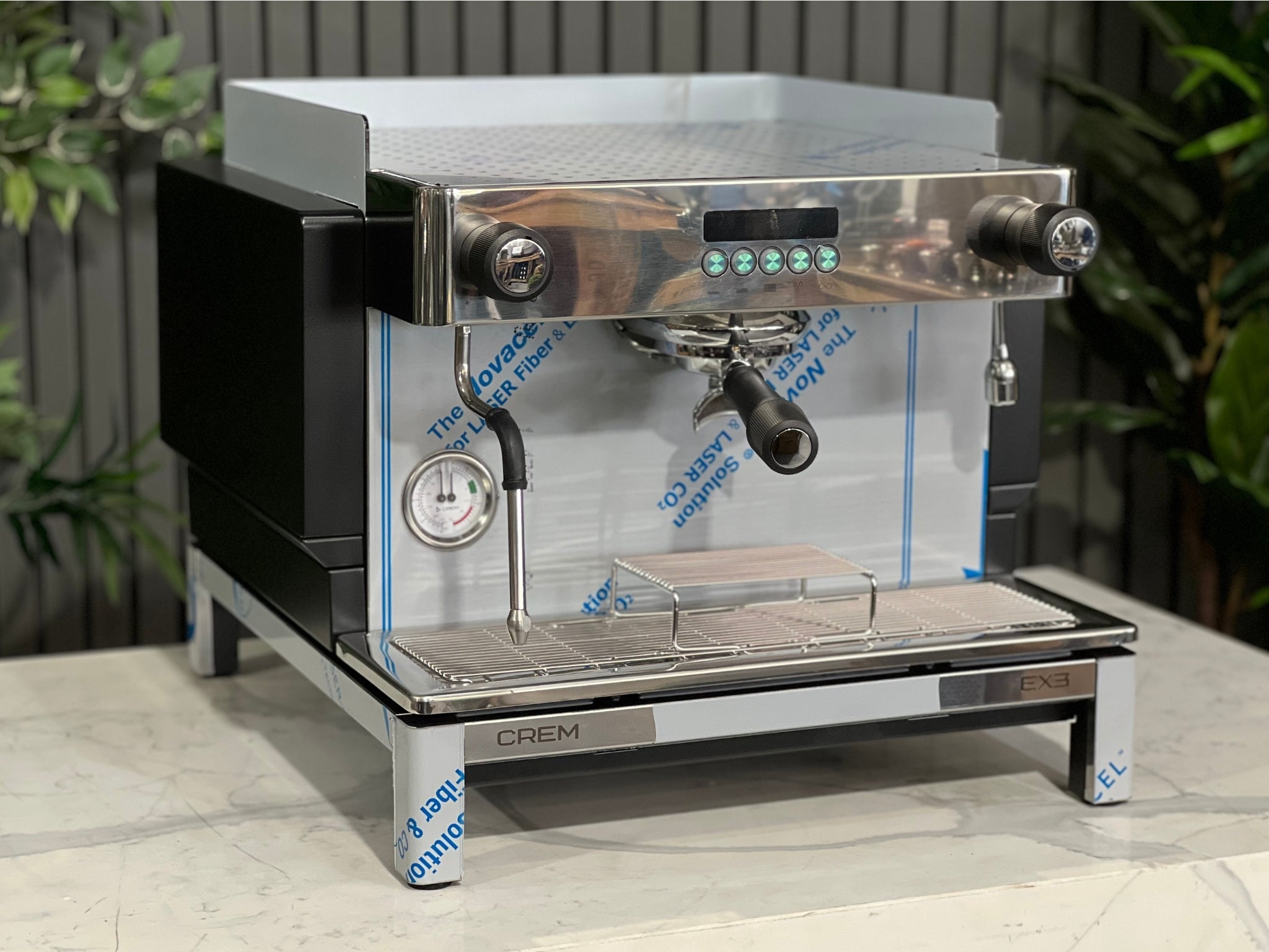 Expobar Crem EX3 Compact 1 Group Coffee Machine Black - New angled view showing the sleek black body, high feet, steam wand, and control panel on a marble bench with greenery in the background. A compact commercial espresso coffee machine built for professional use.