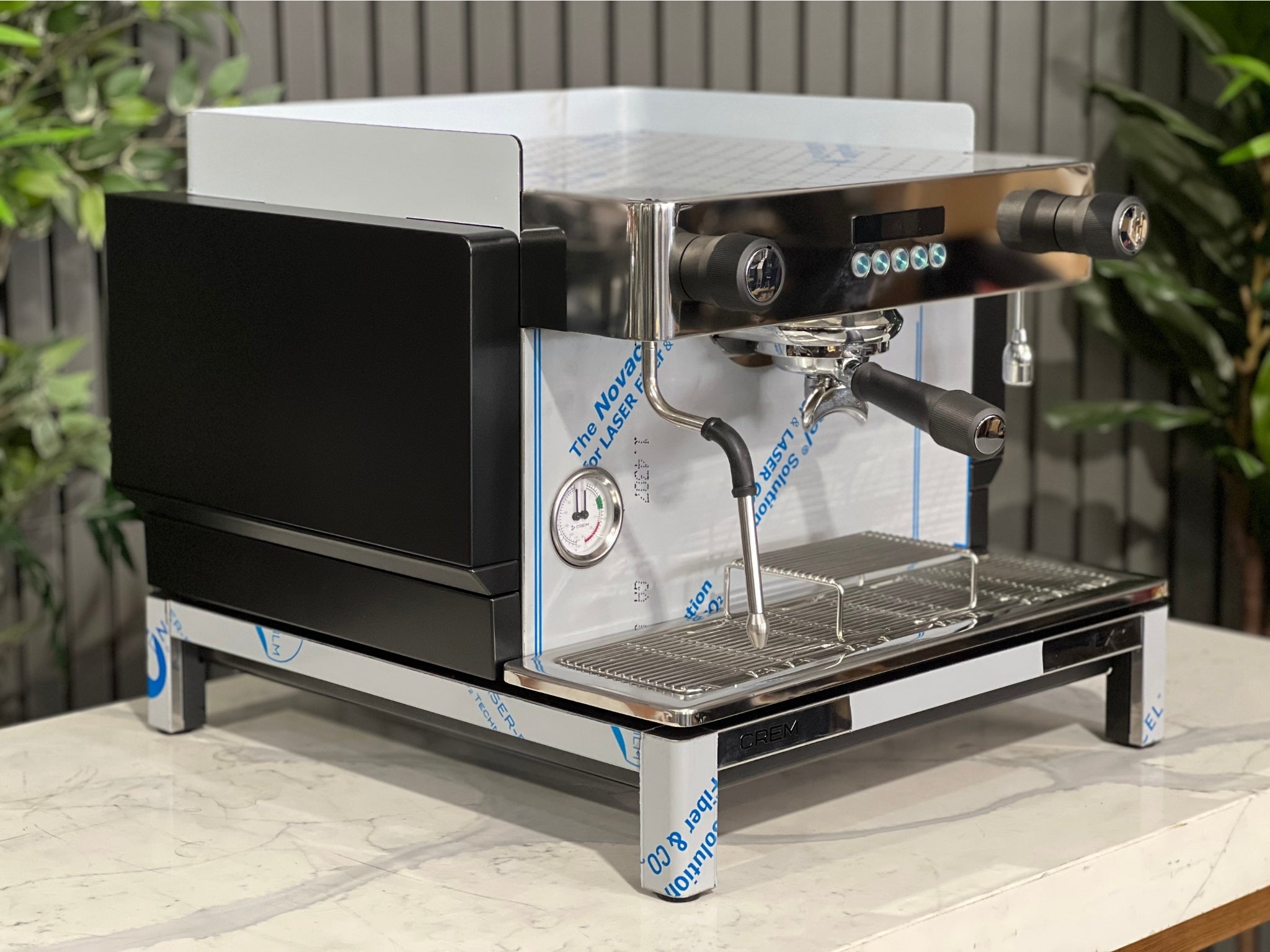 Expobar Crem EX3 Compact 1 Group Coffee Machine Black - New angled view showing the sleek black body, high feet, steam wand, and control panel on a marble bench with greenery in the background. A compact commercial espresso coffee machine built for professional use.