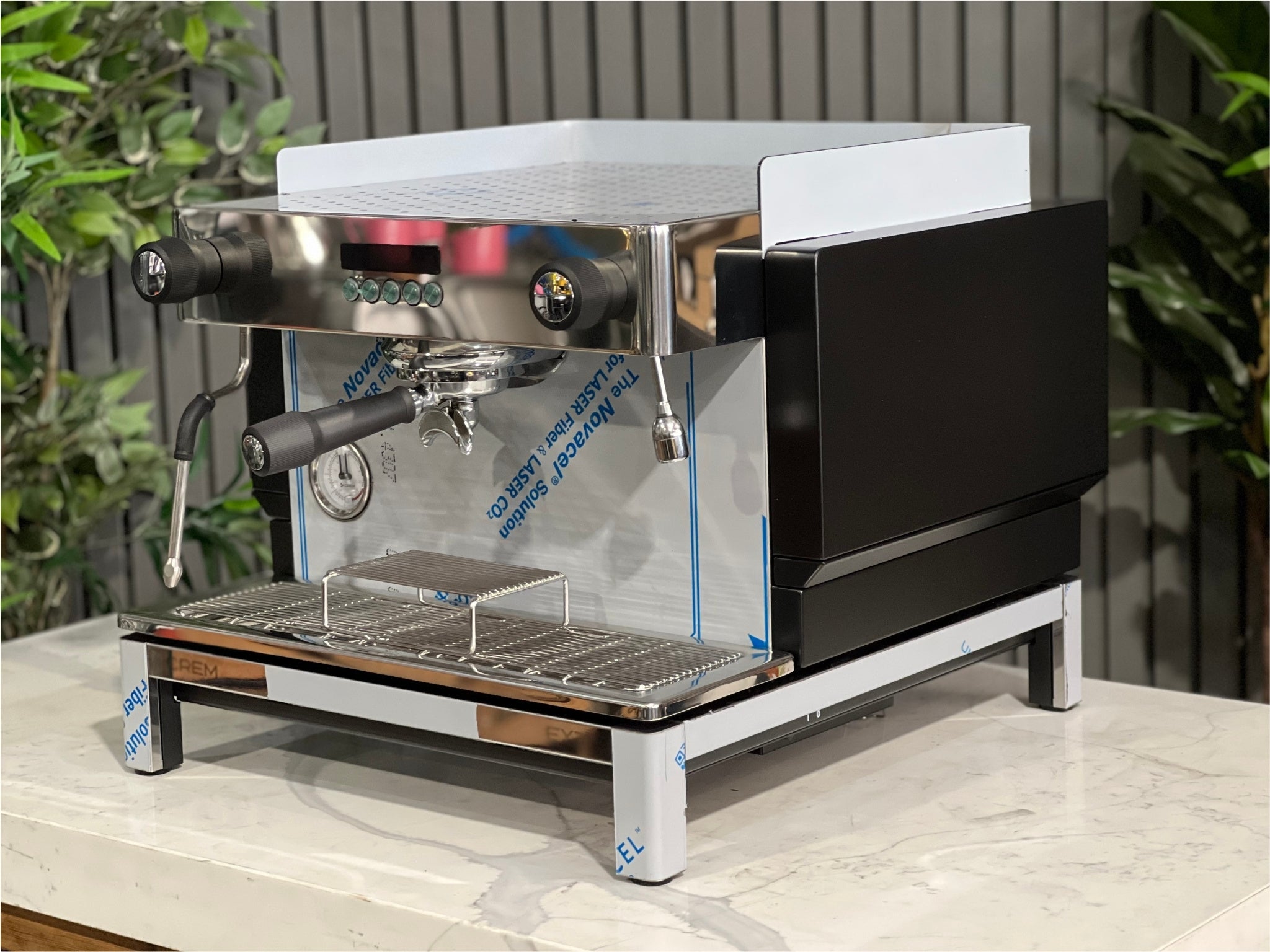 Expobar Crem EX3 Compact 1 Group Coffee Machine Black - New angled view showing the sleek black body, high feet, steam wand, and control panel on a marble bench with greenery in the background. A compact commercial espresso coffee machine built for professional use.