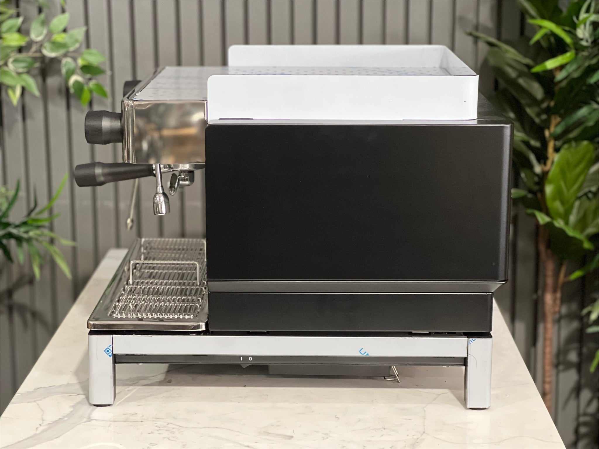 Expobar Crem EX3 Compact 1 Group Coffee Machine Black - New side profile displaying the high feet, steam wand, and elegant side panel detail. An espresso coffee machine made for clean workspace integration, pictured on a marble bench with greenery behind.