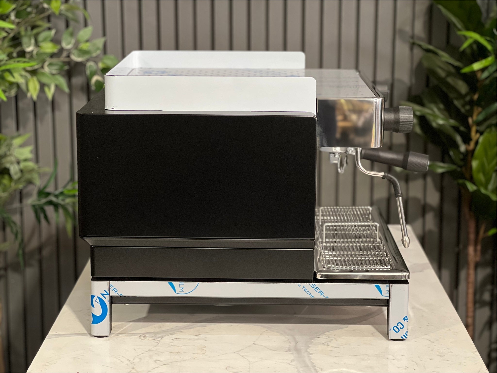 Expobar Crem EX3 Compact 1 Group Coffee Machine Black - New side profile displaying the high feet, steam wand, and elegant side panel detail. An espresso coffee machine made for clean workspace integration, pictured on a marble bench with greenery behind.