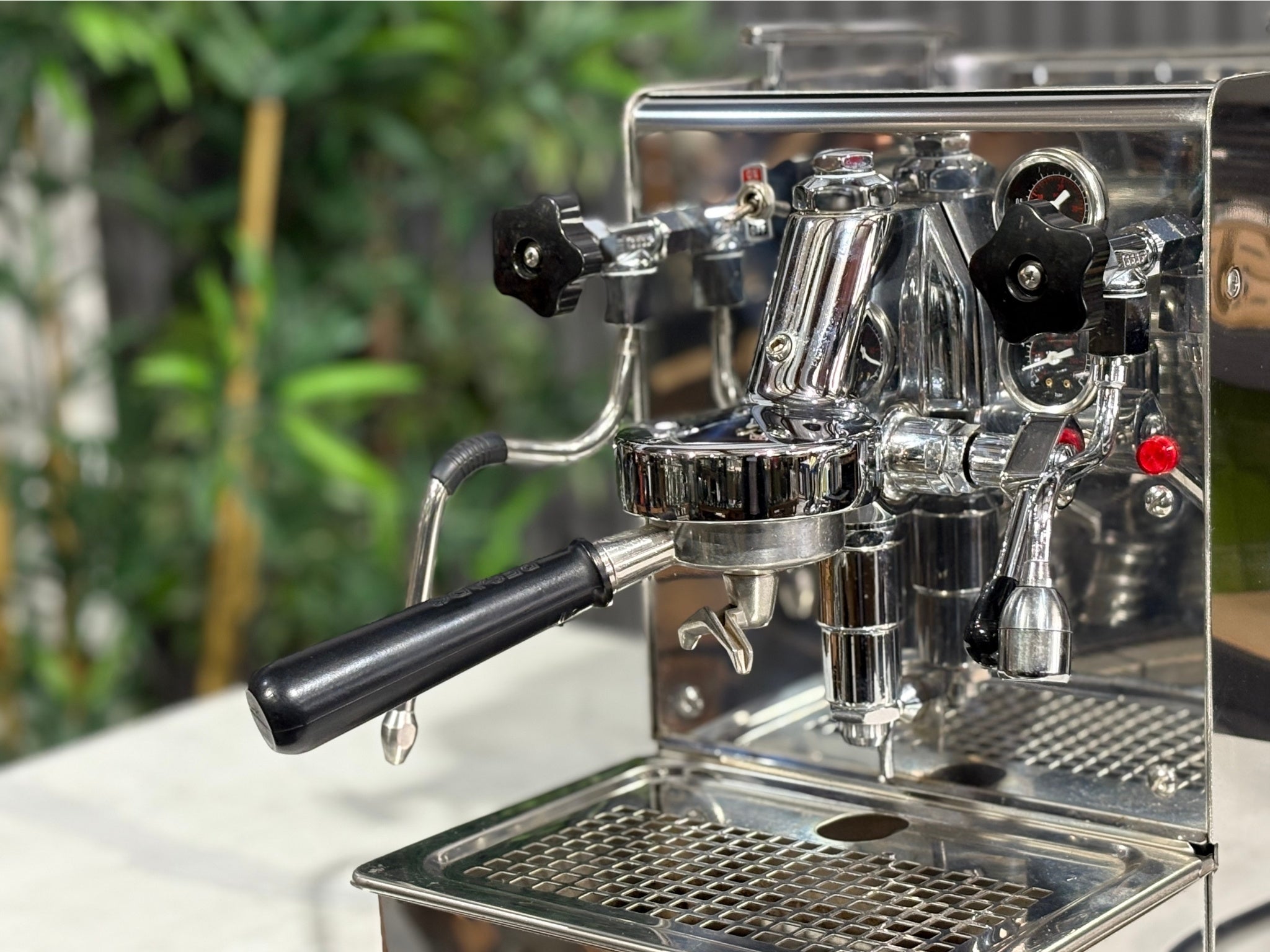 A Expobar Office Leva 1 Group Espresso Coffee Machine with Stainless finish is sitting on a marble bench. There is greenery in the background. The image is a close up image taken from the front of the machine and the machine is sitting on an angle so that you can see the front of the machine, the controls including buttons, group handle and logo.