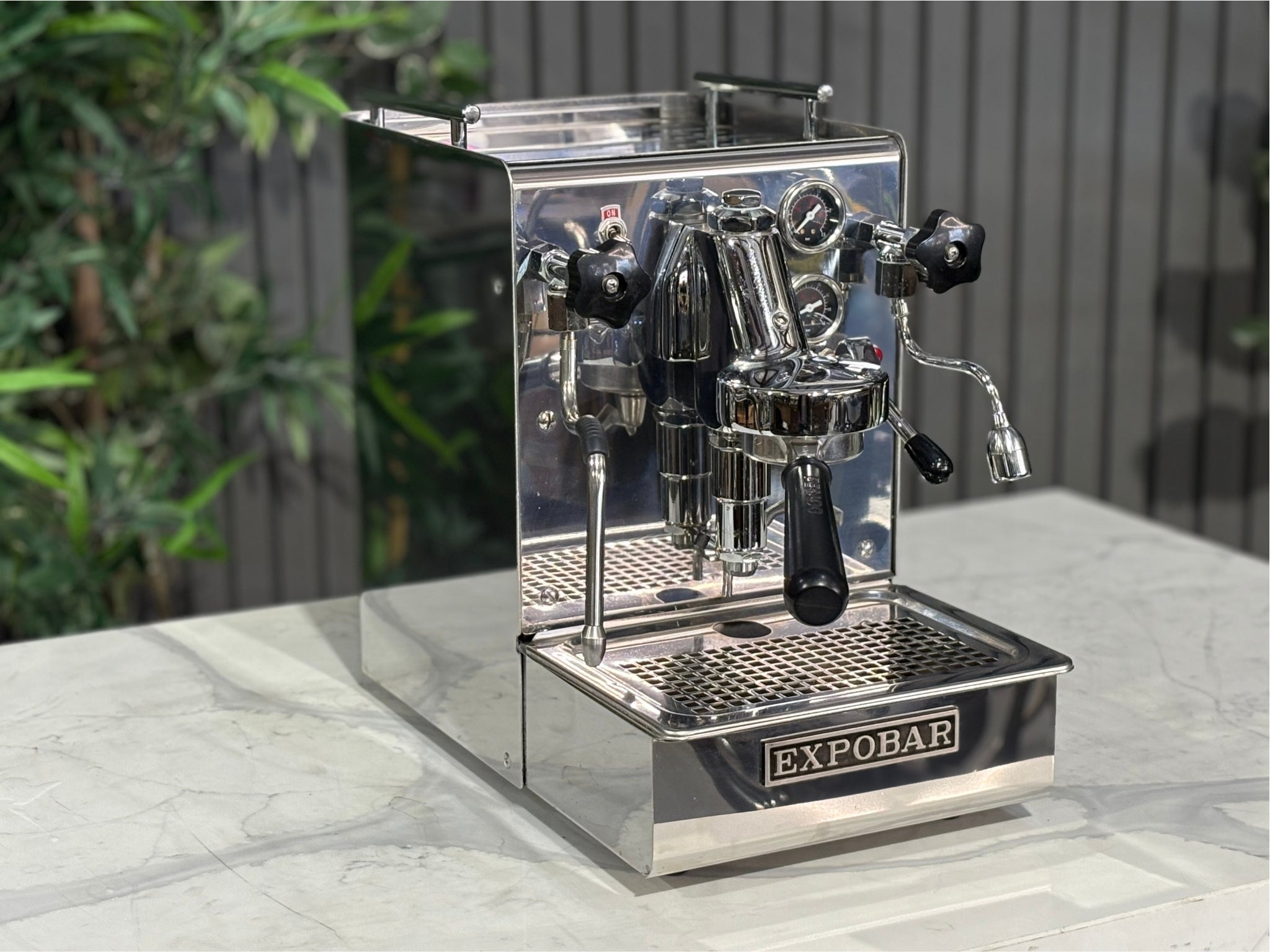 A Expobar Office Leva 1 Group Espresso Coffee Machine with Stainless finish is sitting on a marble bench. There is greenery in the background. The image is taken from the front of the machine and the machine is sitting on an angle so that you can see the front of the machine and the side.