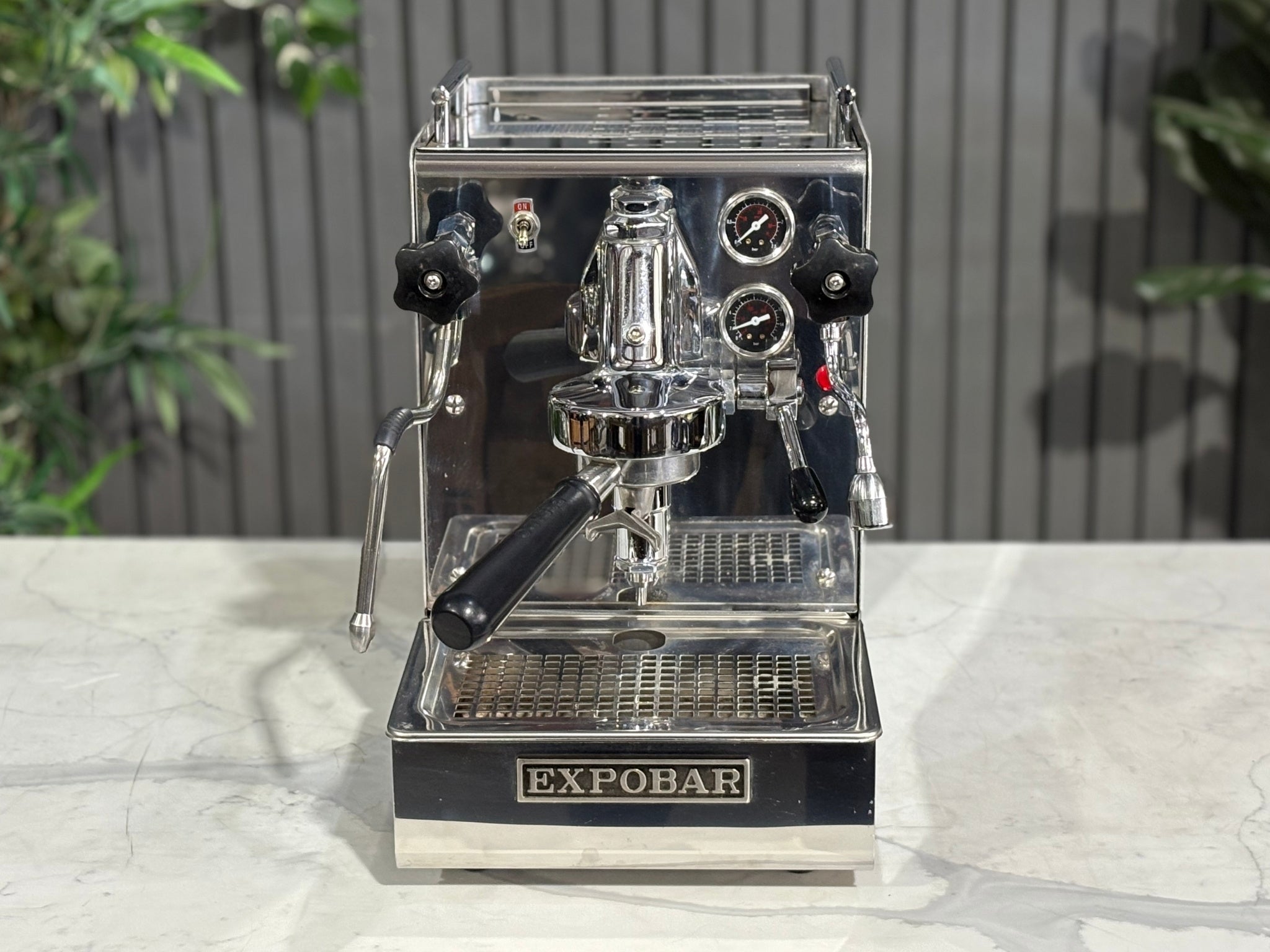 A Expobar Office Leva 1 Group Espresso Coffee Machine with Stainless finish sitting on a marble bench. There is greenery in the background. The photo is taken from a high front angle, clearly showcasing both the front panel and the top of the machine.