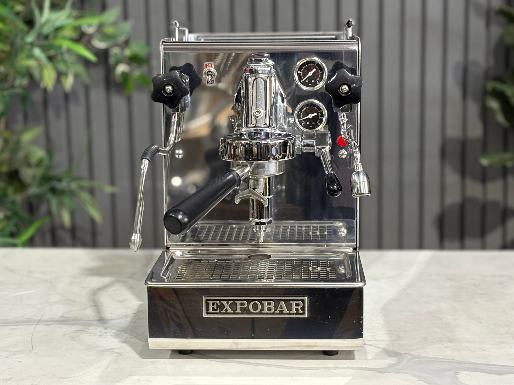A Expobar Office Leva 1 Group Espresso Coffee Machine with Stainless finish sitting on a marble bench. There is greenery in the background. The image is taken from the front of the machine so that you can see the front of the machine, control panel, buttons and group head.