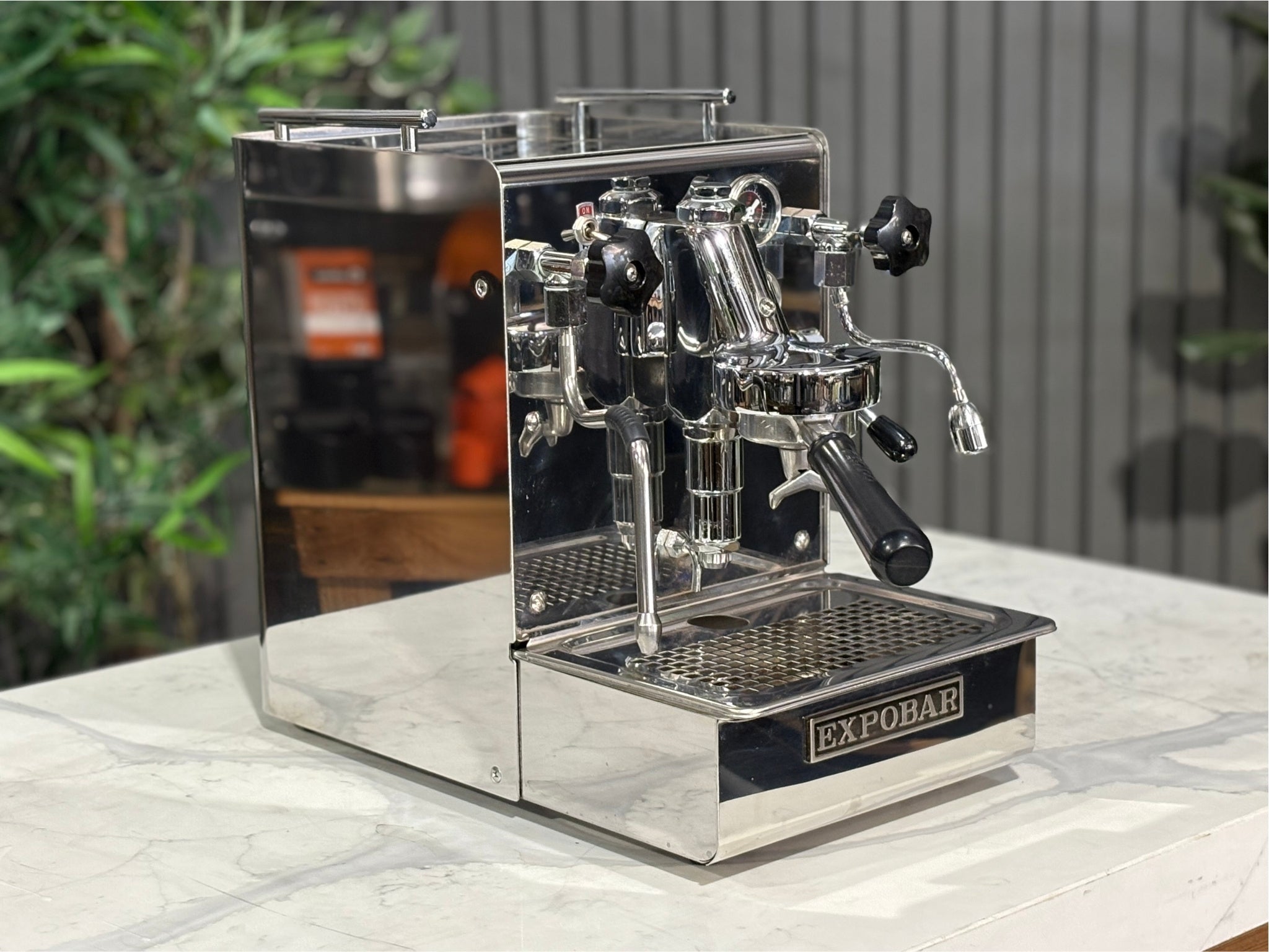 A Expobar Office Leva 1 Group Espresso Coffee Machine with Stainless finish is sitting on a marble bench. There is greenery in the background. The image is taken from the front of the machine and the machine is sitting on an angle so that you can see the front of the machine and the side.