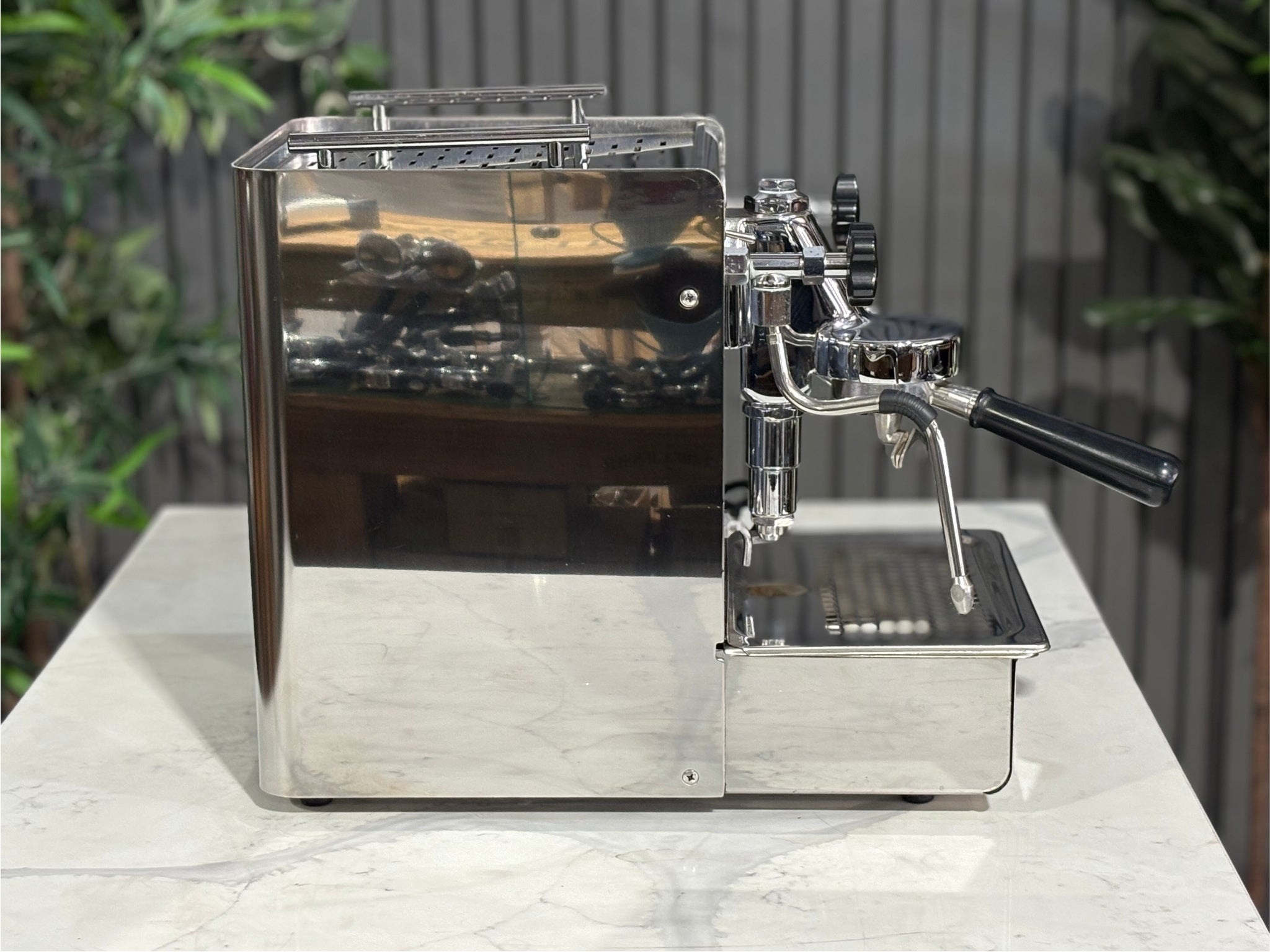 A Expobar Office Leva 1 Group Espresso Coffee Machine with Stainless finish sitting on a marble bench. There is greenery in the background. The image is taken from the side of the machine.