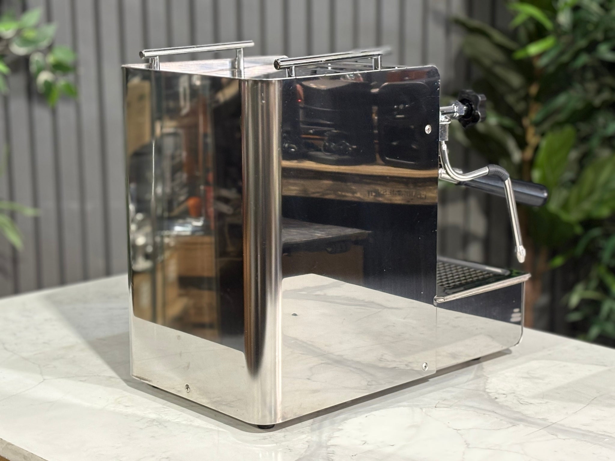 A Expobar Office Leva 1 Group Espresso Coffee Machine with Stainless finish sitting on a marble bench. There is greenery in the background. The image is taken from behind the machine and the machine is sitting on an angle so that you can see the back of the machine and the side.