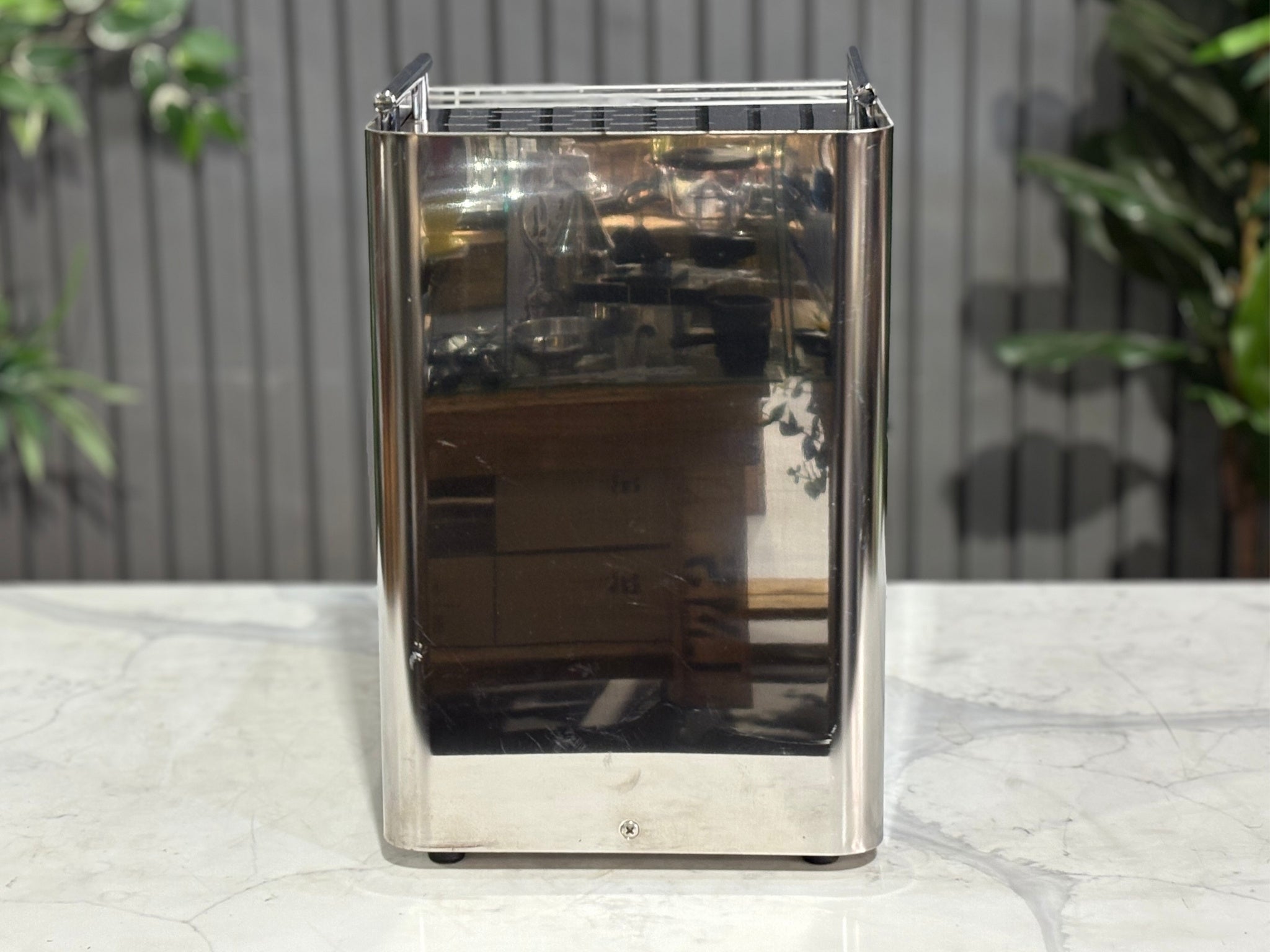 A Expobar Office Leva 1 Group Espresso Coffee Machine with Stainless finish is sitting on a marble bench. There is greenery in the background. The image is taken from behind the machine so the back is visible.