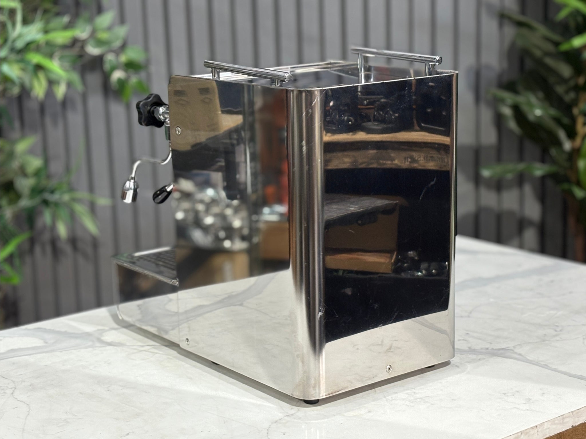 A Expobar Office Leva 1 Group Espresso Coffee Machine with Stainless finish sitting on a marble bench. There is greenery in the background. The image is taken from behind the machine and the machine is sitting on an angle so that you can see the back of the machine and the side.