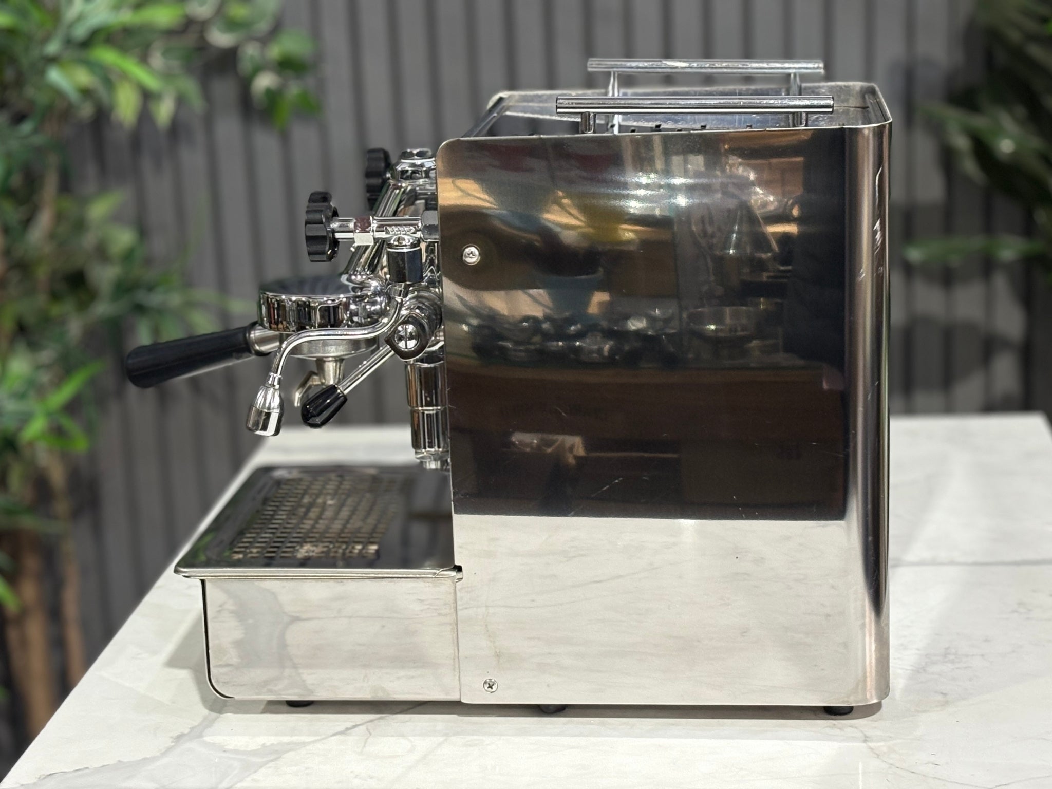 A Expobar Office Leva 1 Group Espresso Coffee Machine with Stainless finish sitting on a marble bench. There is greenery in the background. The image is taken from the side of the machine.