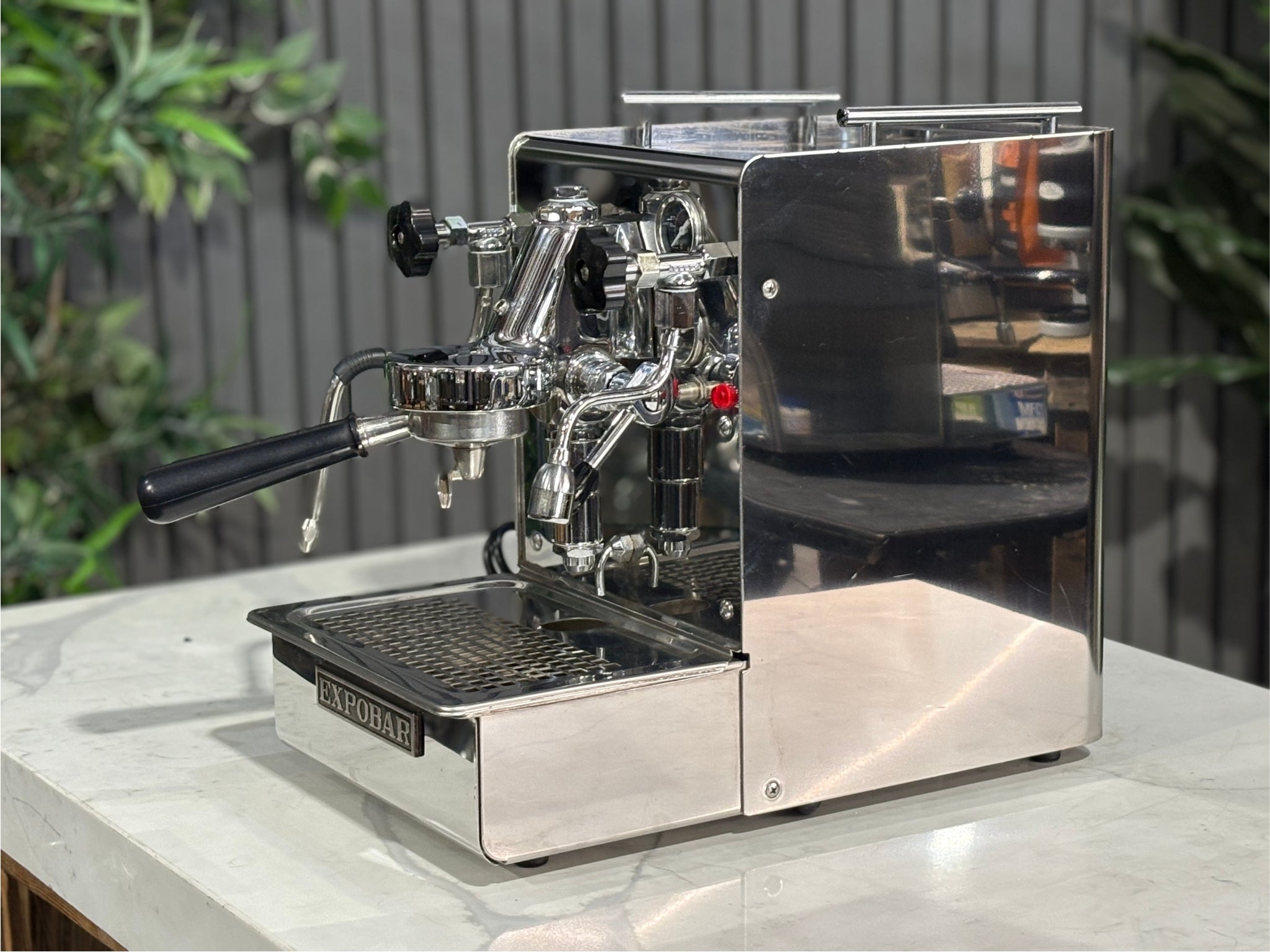 A Expobar Office Leva 1 Group Espresso Coffee Machine with Stainless finish is sitting on a marble bench. There is greenery in the background. The image is taken from the front of the machine and the machine is sitting on an angle so that you can see the front of the machine and the side.