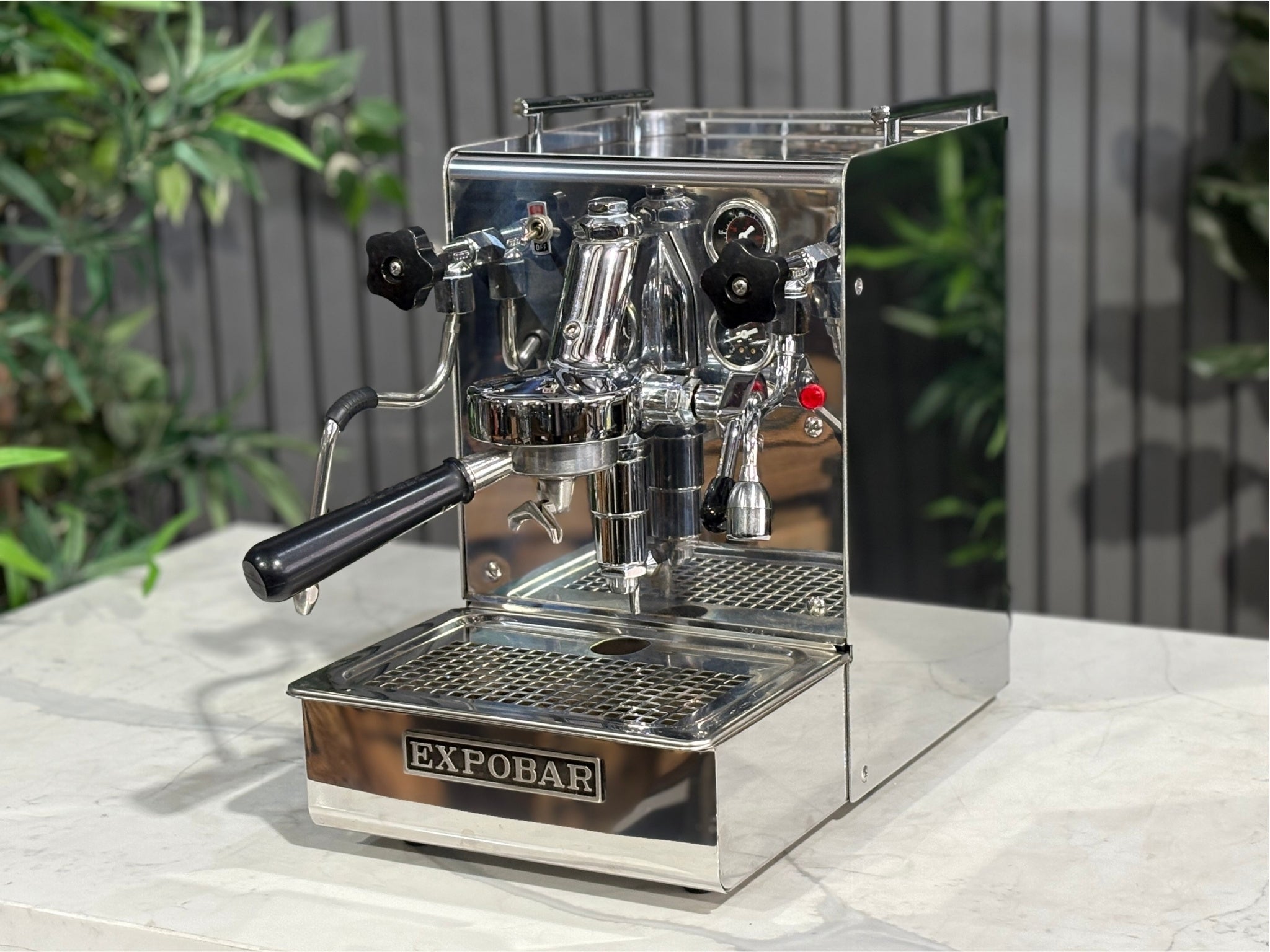 A Expobar Office Leva 1 Group Espresso Coffee Machine with Stainless finish is sitting on a marble bench. There is greenery in the background. The image is taken from the front of the machine and the machine is sitting on an angle so that you can see the front of the machine and the side.