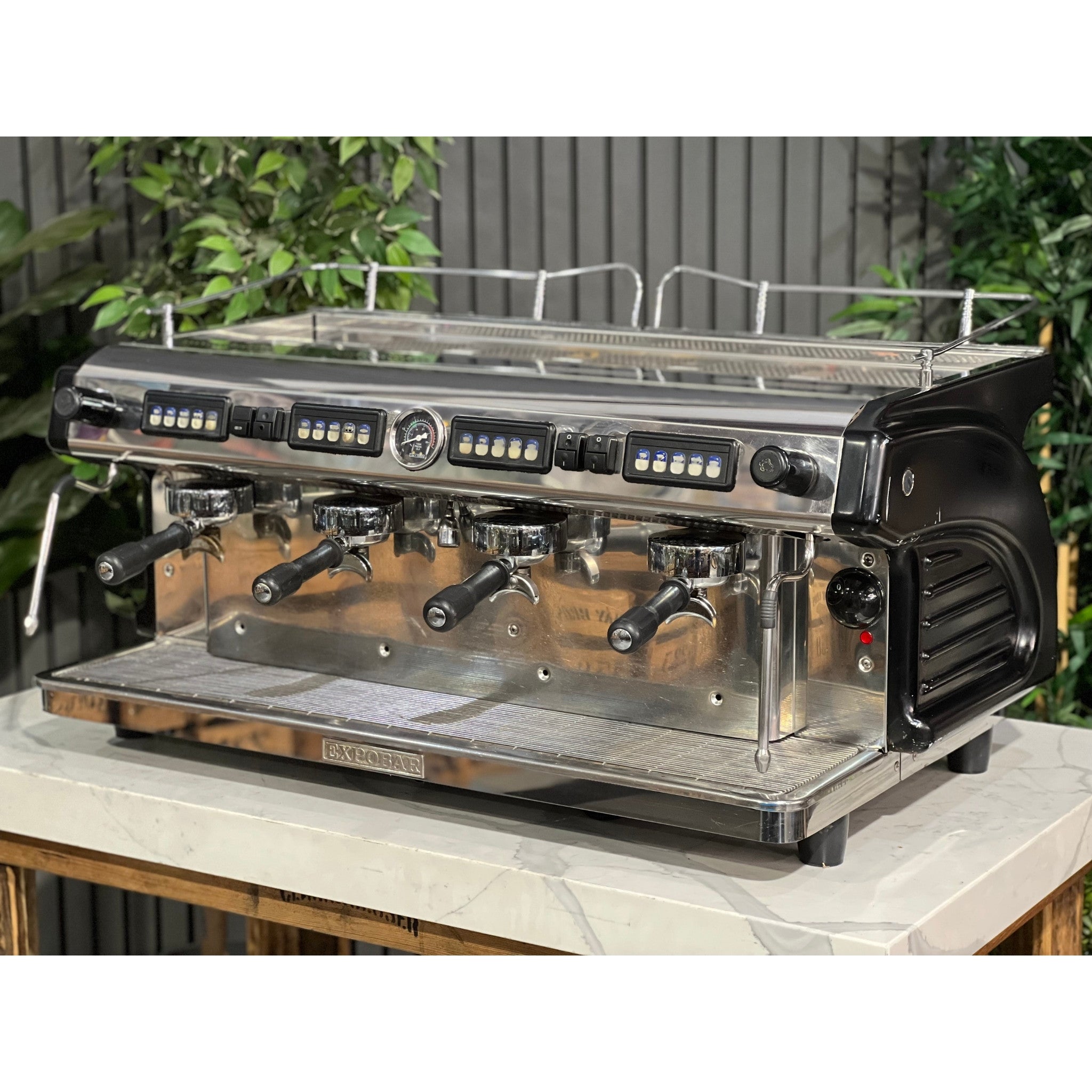 Expobar Ruggero 4 Group Black Espresso Coffee Machine in Stainless steel is sitting on a wooden bench. There is greenery in the background. The image is taken from the front of the machine and the machine is sitting on an angle so that you can see the front of the machine and the side