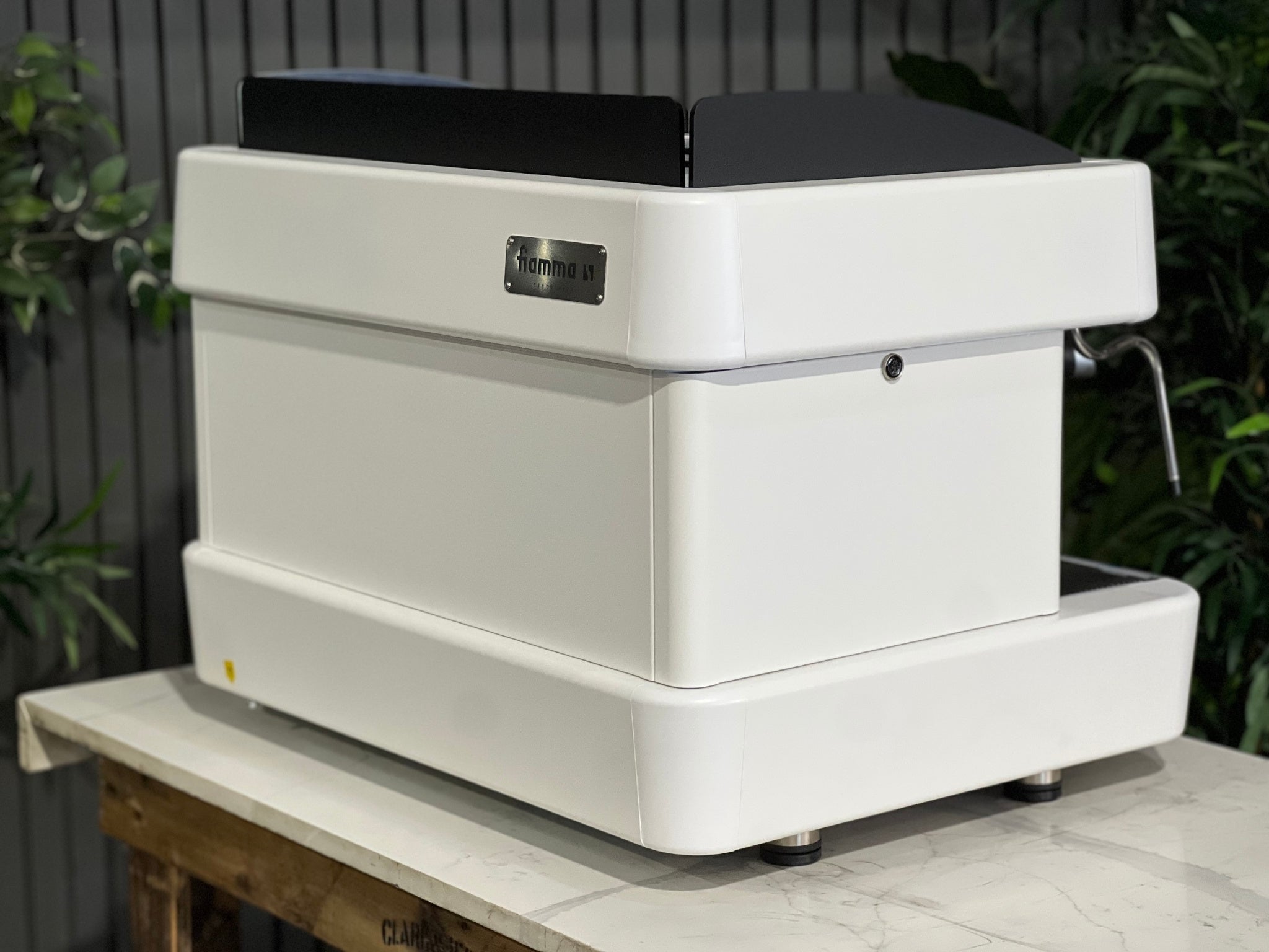 A brand new Fiamma Quadrant 2 DSP 2 Group Coffee Machine with White finish 
sitting on a marble bench. There is greenery in the background. The image is taken from behind the machine and the machine is sitting on an angle so that you can see the back of the machine and the side.