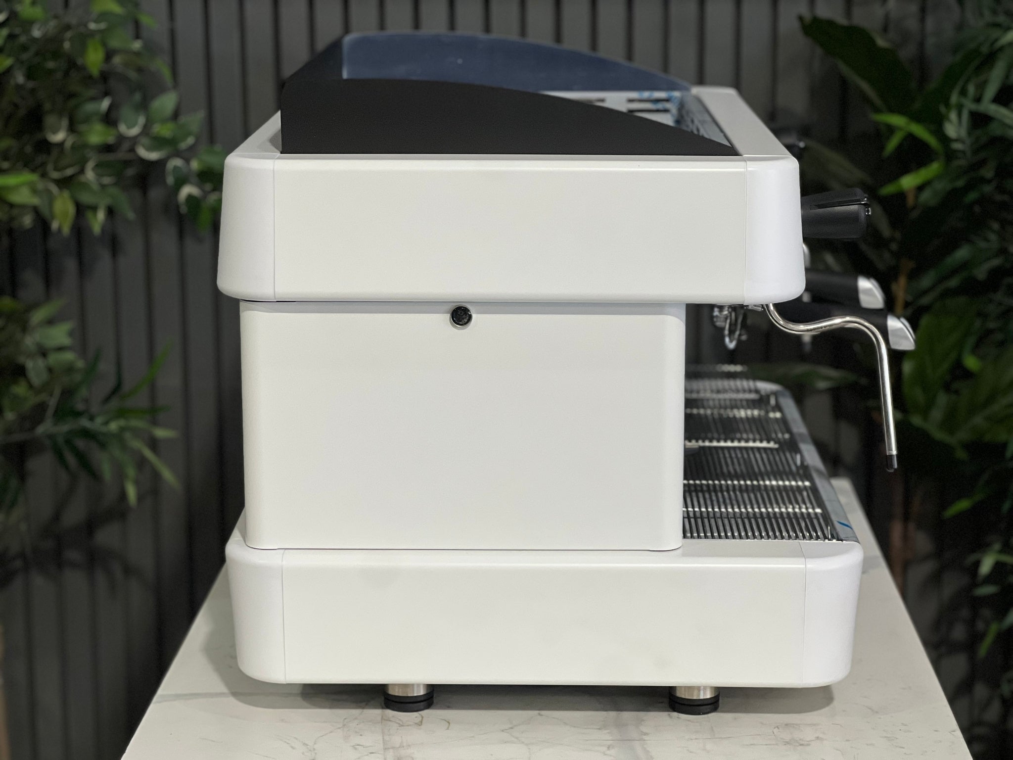A brand new Fiamma Quadrant 2 DSP 2 Group Coffee Machine with White finish  sitting on a marble bench. There is greenery in the background. The image is taken from the side of the machine.