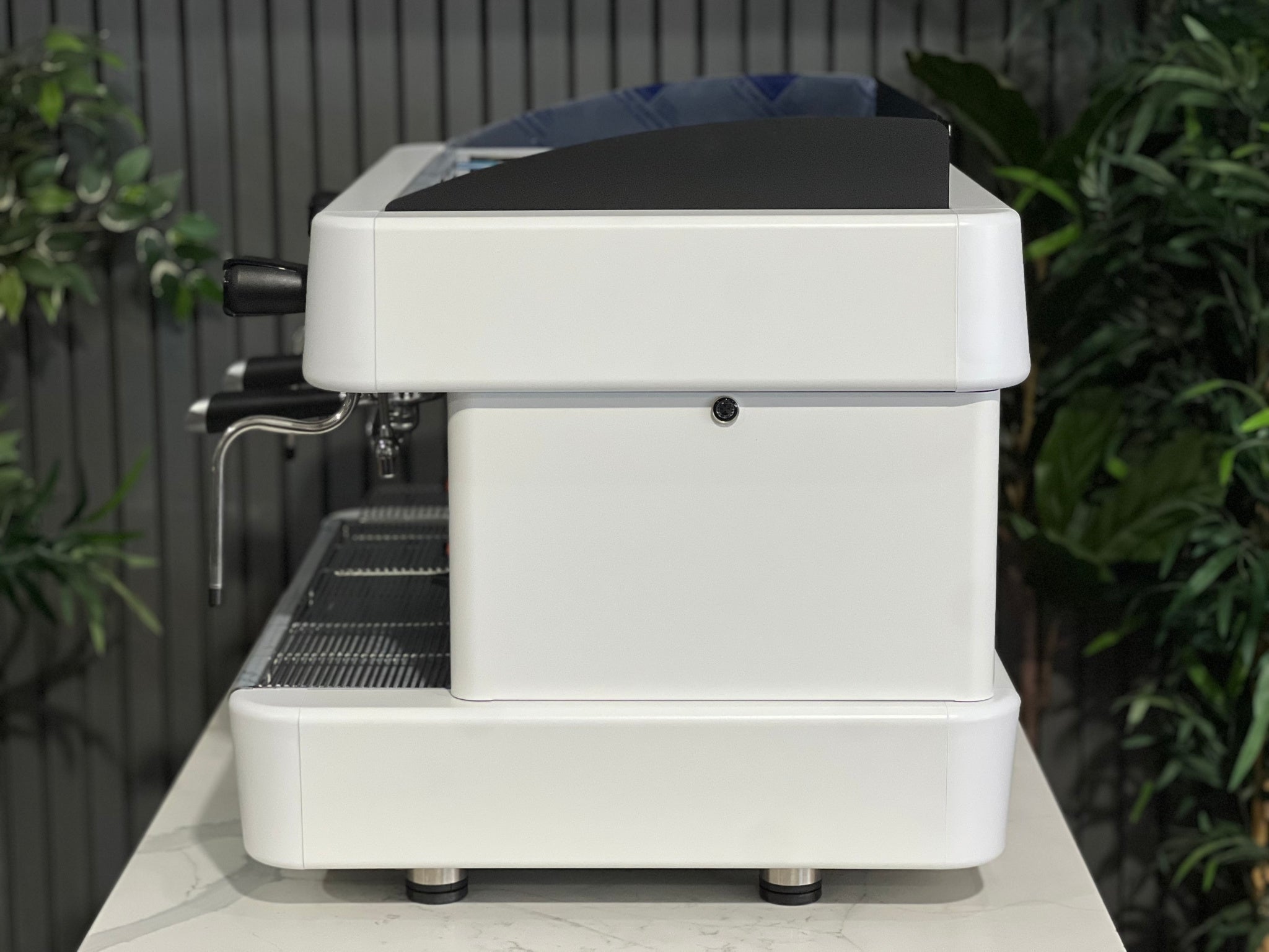 A brand new Fiamma Quadrant 2 DSP 2 Group Coffee Machine with White finish sitting on a marble bench. There is greenery in the background. The image is taken from the side of the machine.