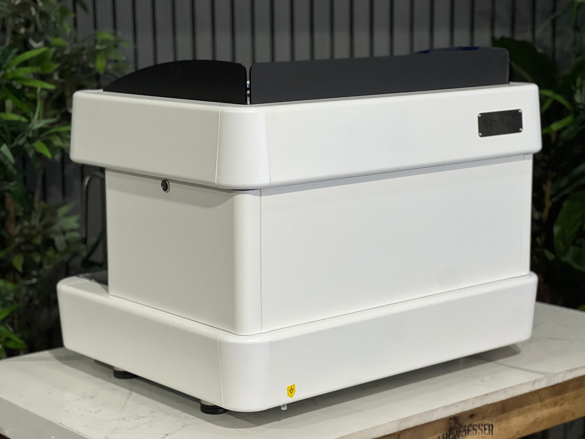 A brand new Fiamma Quadrant 2 DSP 2 Group Coffee Machine with White finish 
sitting on a marble bench. There is greenery in the background. The image is taken from behind the machine and the machine is sitting on an angle so that you can see the back of the machine and the side.