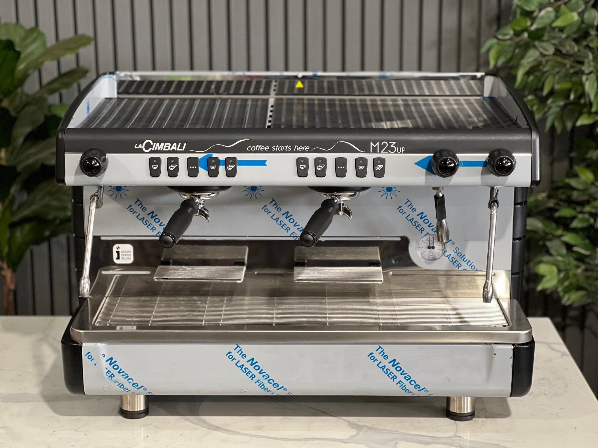 A brand new La Cimbali M23 UP 2 Group Coffee Machine with Black finish sitting on a marble bench. There is greenery in the background. The image is taken from the front of the machine so that you can see the front of the machine, control panel, buttons and group heads.