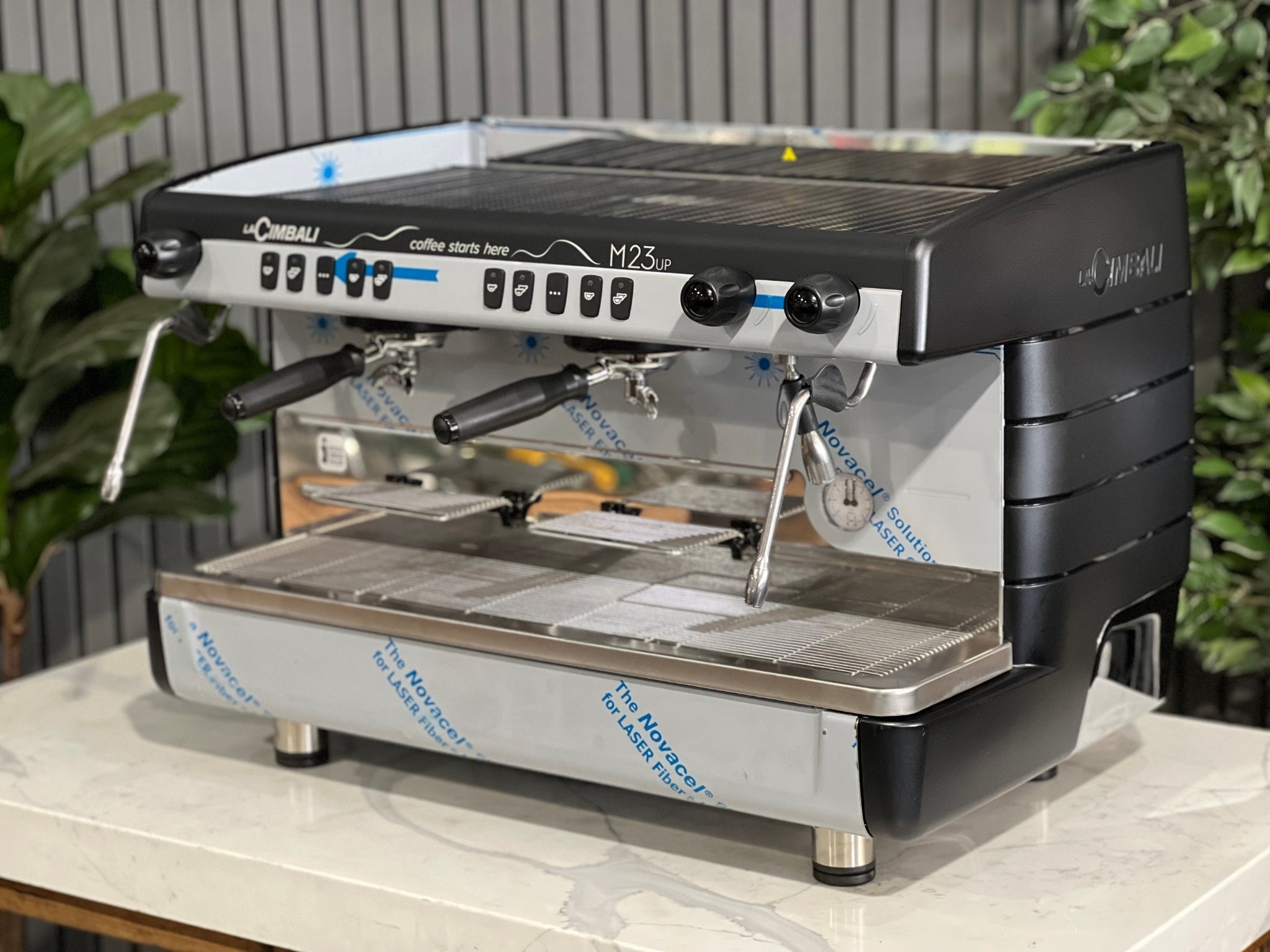 A brand new La Cimbali M23 UP 2 Group Coffee Machine with Black finish is sitting on a marble bench. There is greenery in the background. The image is taken from the front of the machine and the machine is sitting on an angle so that you can see the front of the machine and the side.