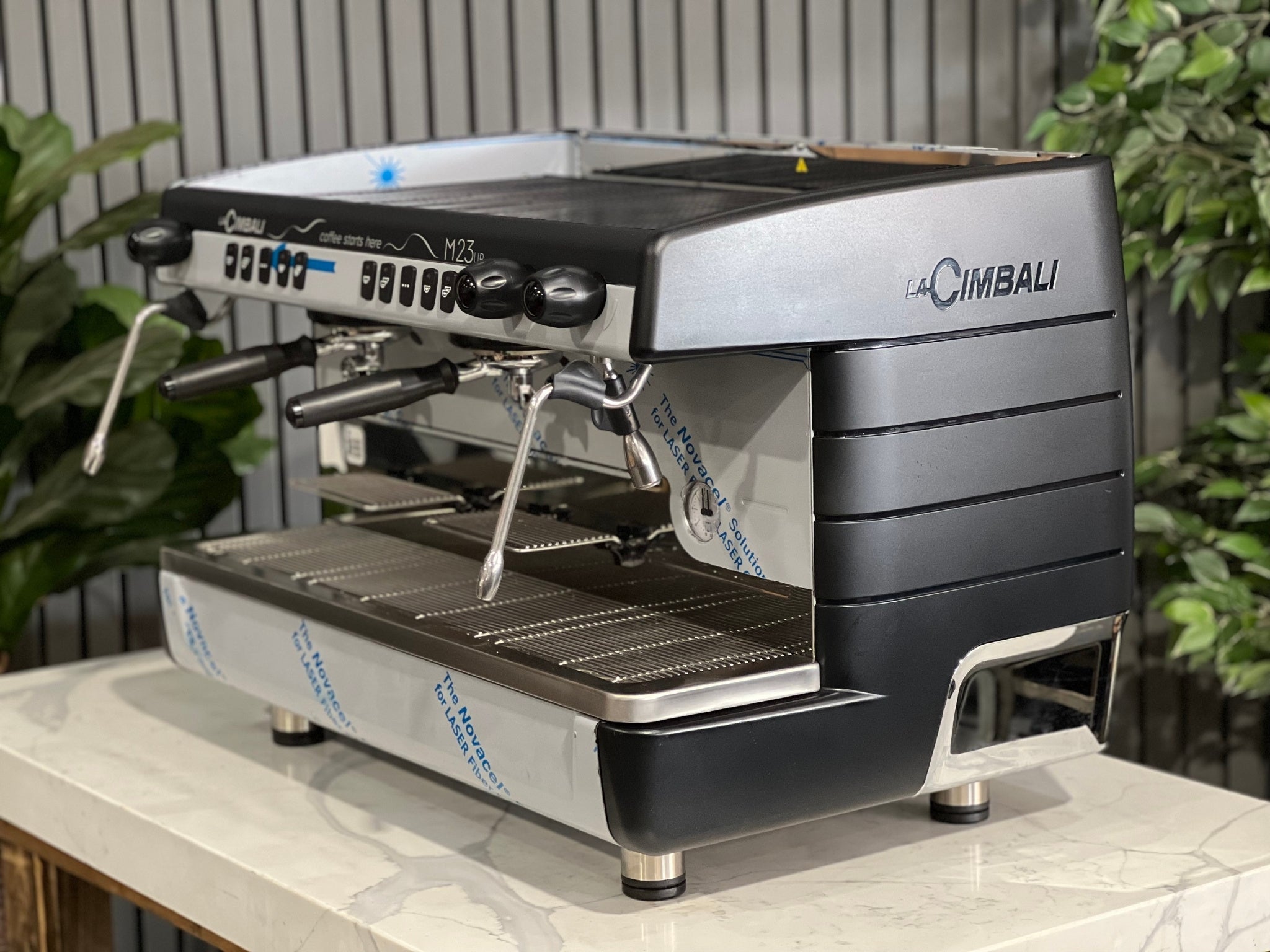 A brand new La Cimbali M23 UP 2 Group Coffee Machine with Black finish is sitting on a marble bench. There is greenery in the background. The image is taken from the front of the machine and the machine is sitting on an angle so that you can see the front of the machine and the side.
