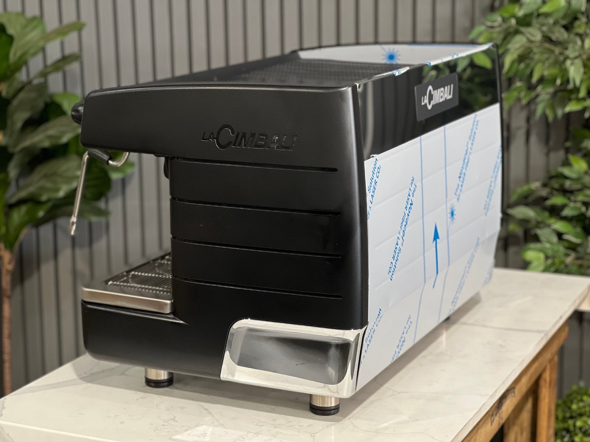 A brand new La Cimbali M23 UP 2 Group Coffee Machine with Black finish sitting on a marble bench. There is greenery in the background. The image is taken from behind the machine and the machine is sitting on an angle so that you can see the back of the machine and the side.